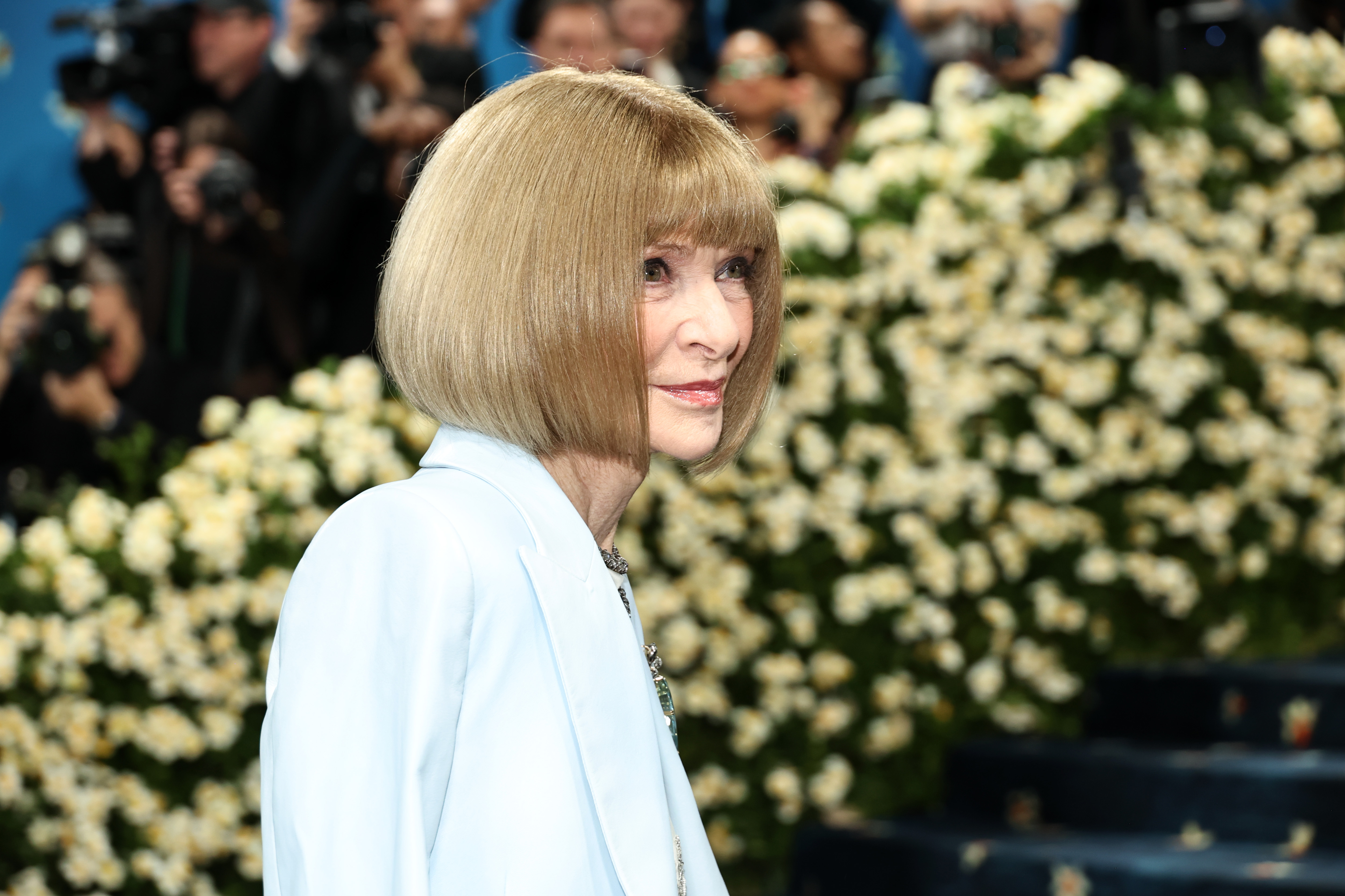 Anna Wintour glances slightly over her shoulder on the Met Gala carpet, surrounded by floral arrangements, as she attends the 2025 celebration of "Superfine: Tailoring Black Style" at the Metropolitan Museum of Art.