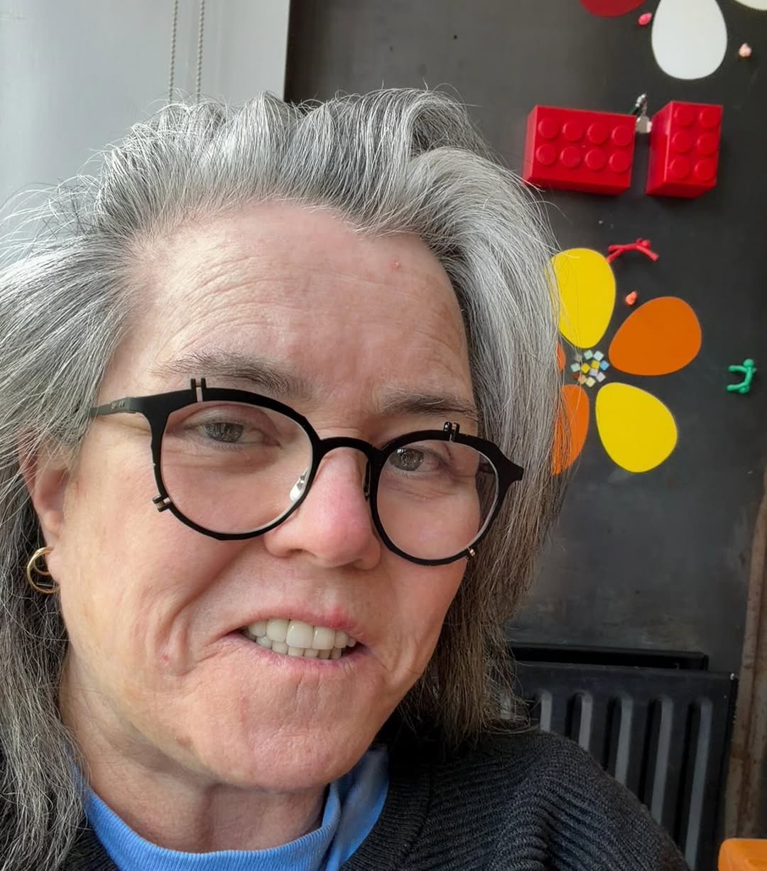 Rosie O'Donnell smiles softly in a casual selfie beside colorful wall art and bright red toy blocks, wearing round glasses and layered clothing in a cozy home setting. | Source: Instagram/rosie
