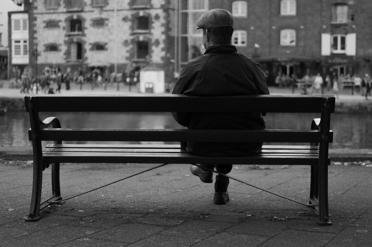 A black-and-white image of a man sitting on a bench looking at the view in front of him | Photo: Pixabay