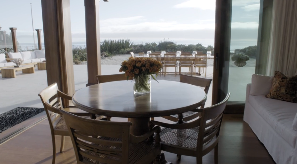 A round wooden dining table topped with a vase of golden flowers sits inside an sunlit breakfast nook surrounded by woven-back chairs. Pocketing glass doors frame direct views of the outdoor dining terrace, coastal dune vegetation, and the ocean beyond. | Source: YouTube/Architectural Digest