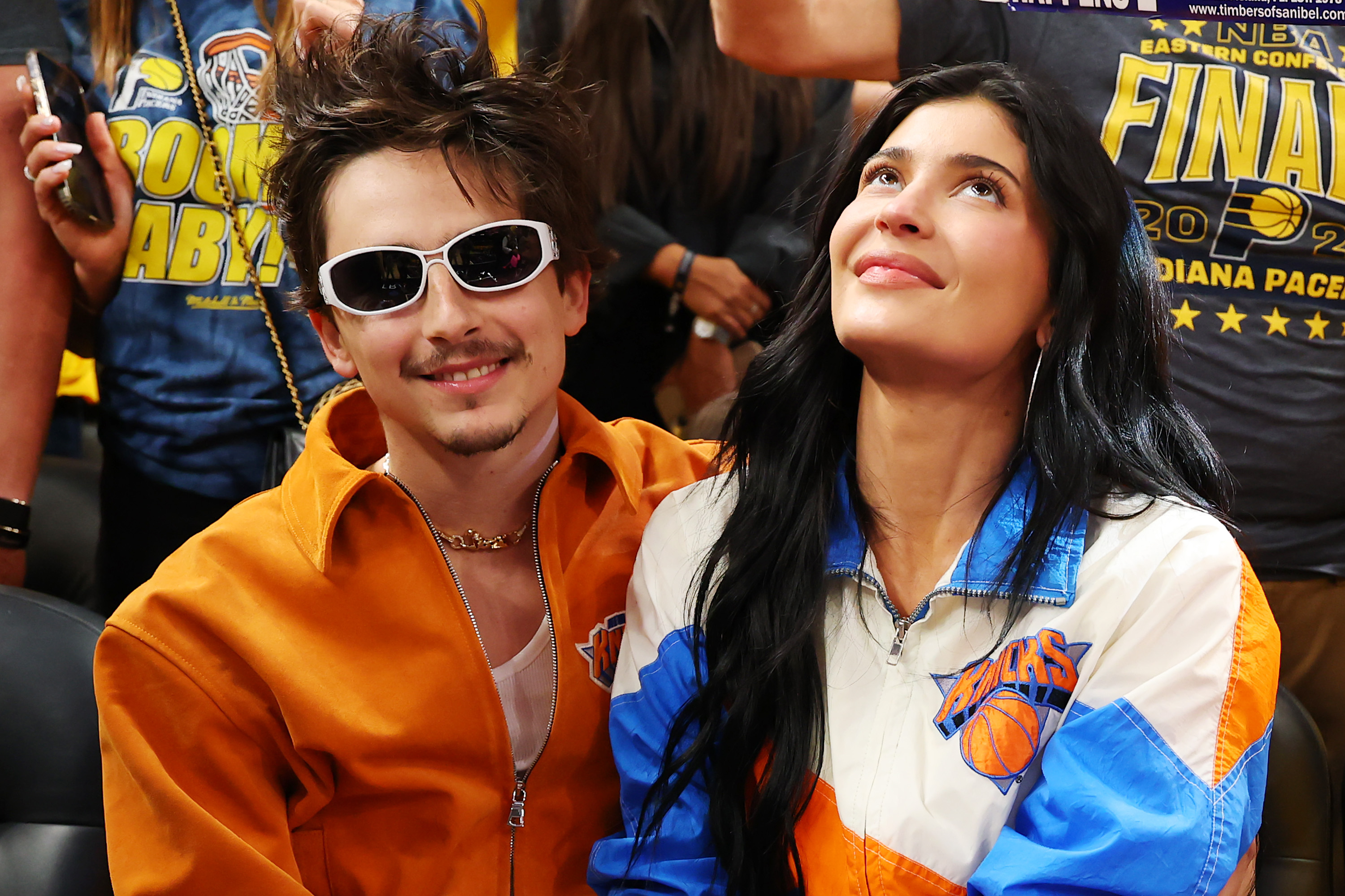 Timothée Chalamet, in an orange outfit with his curly hair loose and messy, playfully smiled at the camera in white-rimmed sunglasses while attending Game Six of the Eastern Conference Finals with Kylie Jenner on May 31, 2025, at Gainbridge Fieldhouse in Indianapolis, Indiana.