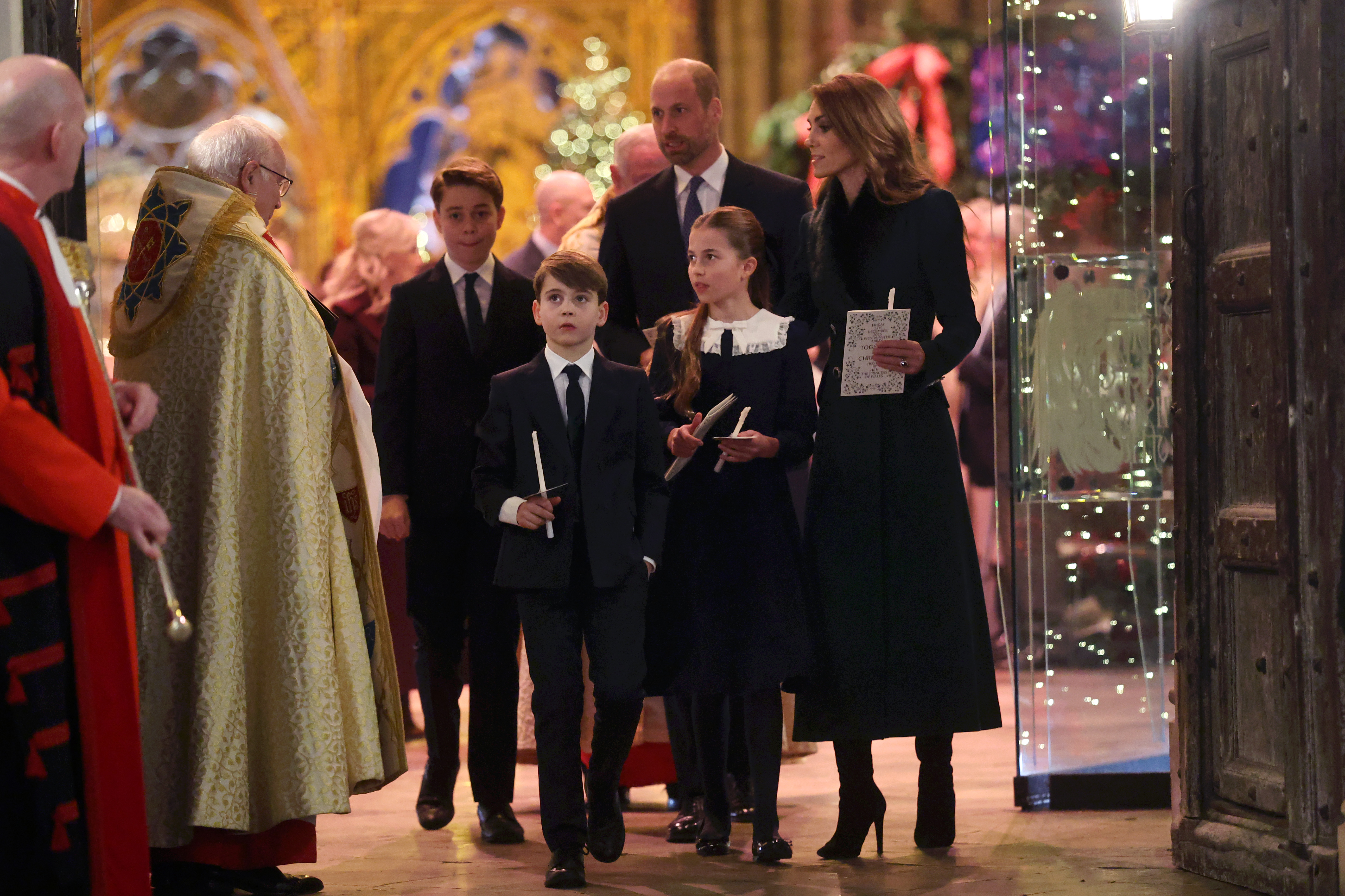 Inside the Abbey, the Wales family paused momentarily with candles and programmes in hand. Prince Louis led the way with quiet confidence, while Princess Charlotte stood attentively between her parents. Catherine, Princess of Wales, exuded elegance as they were welcomed into the softly lit space.