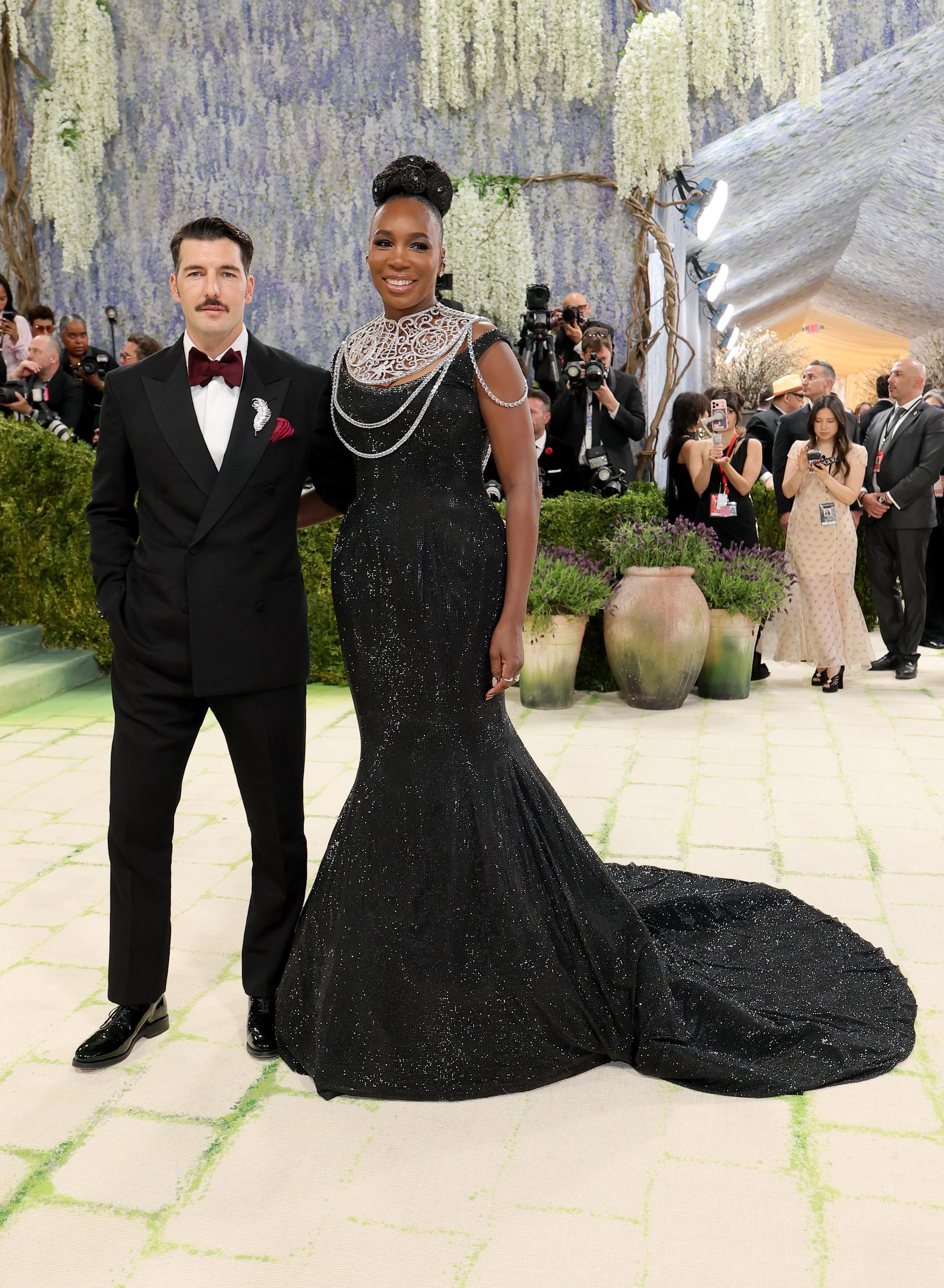 Andrea Preti and Venus Williams pose together at the 2026 Met Gala, with Preti in a black tuxedo and burgundy bow tie and Williams in a dramatic black gown with a sweeping train.