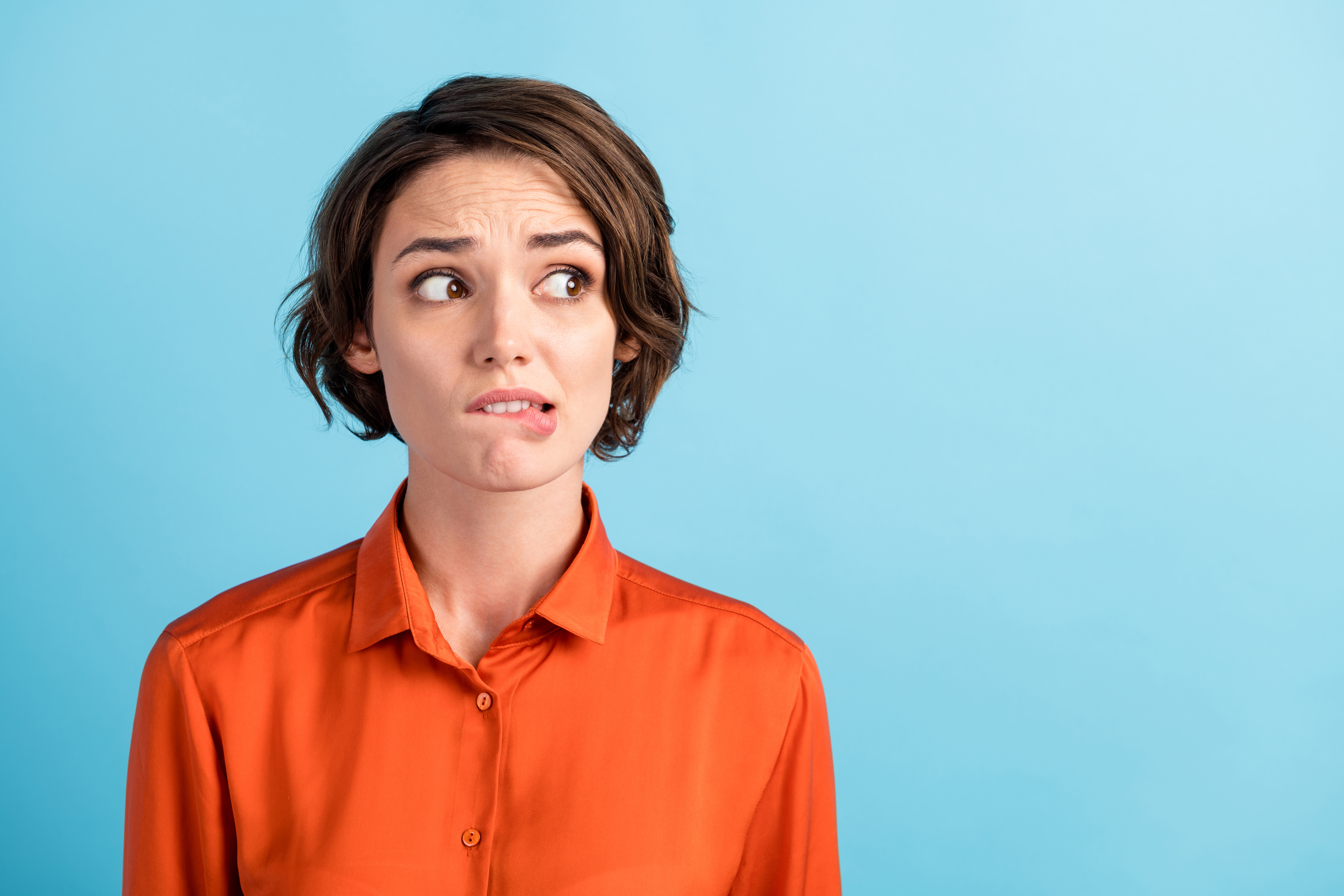 Closeup photo of sad depressed displeased lady horrified facial expression made huge big mistake feel guilty look side empty space bite lips wear orange shirt isolated blue color background | Source: Getty Images