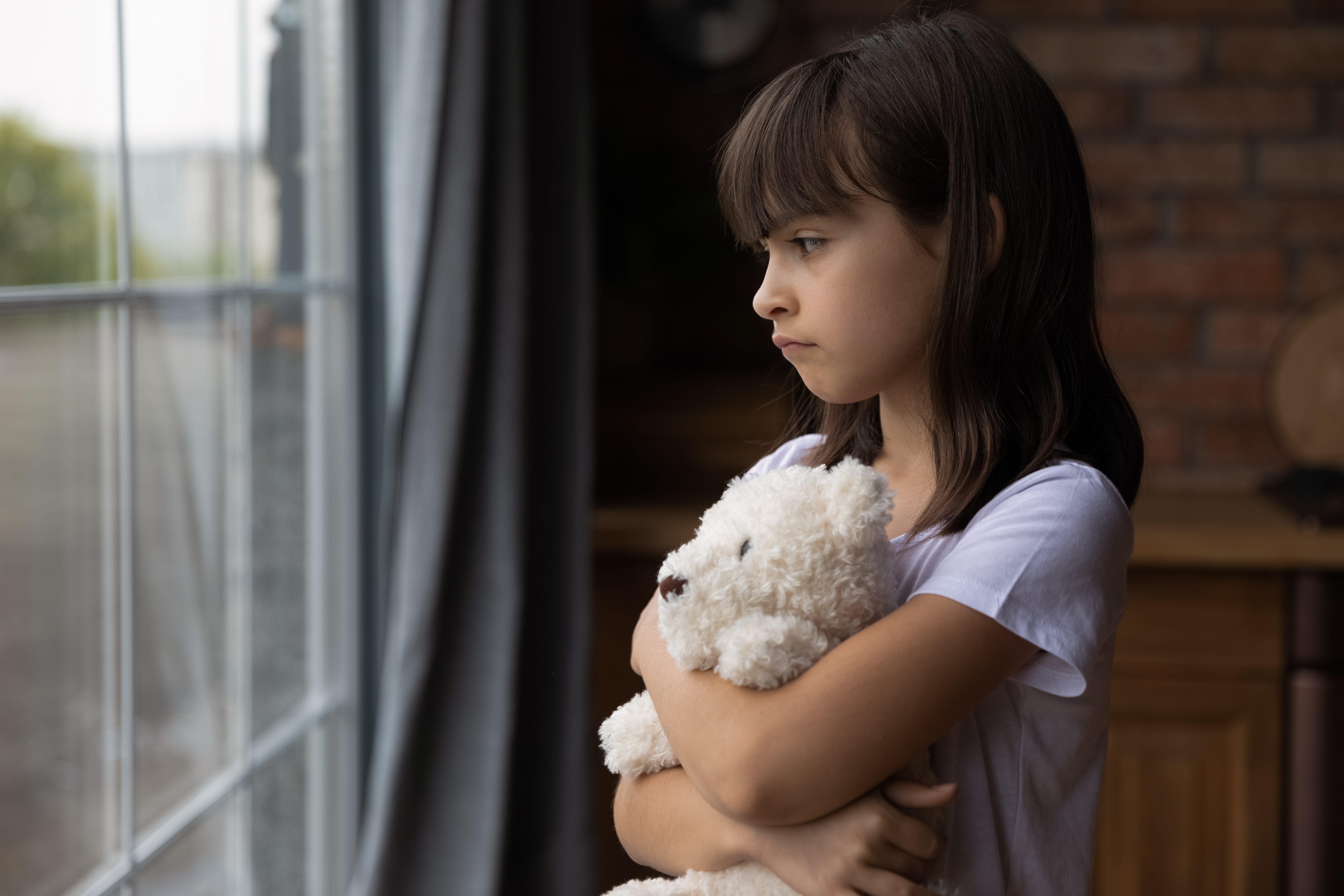 A girl looking outside a window while hugging a toy | Source: Shutterstock