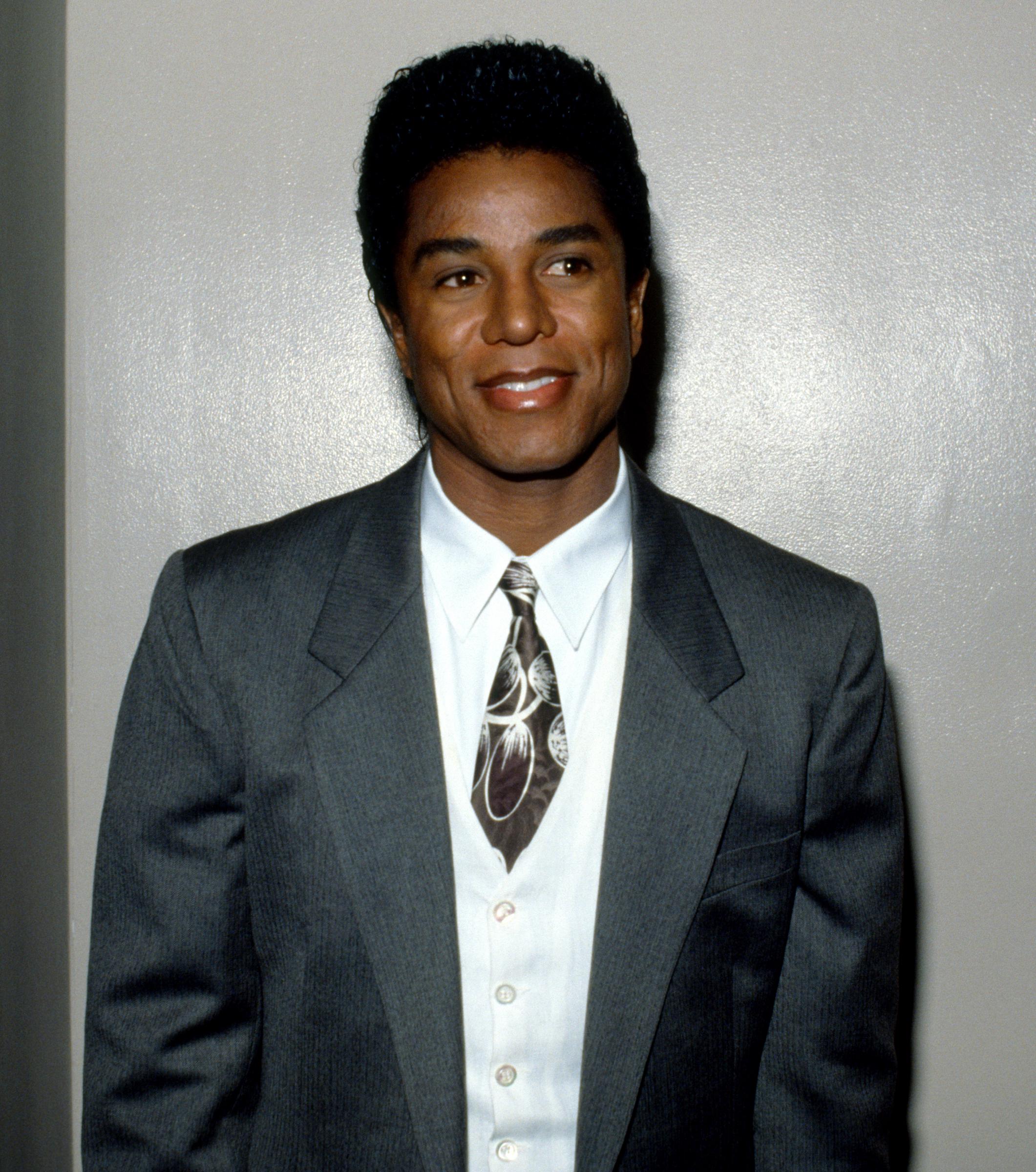 Dressed in a tailored gray suit with a patterned tie, Jermaine Jackson poses with composed confidence, reflecting a more refined, late-1980s image. His subtle smile and clean-cut styling mark a shift toward sophistication while maintaining his signature charisma.