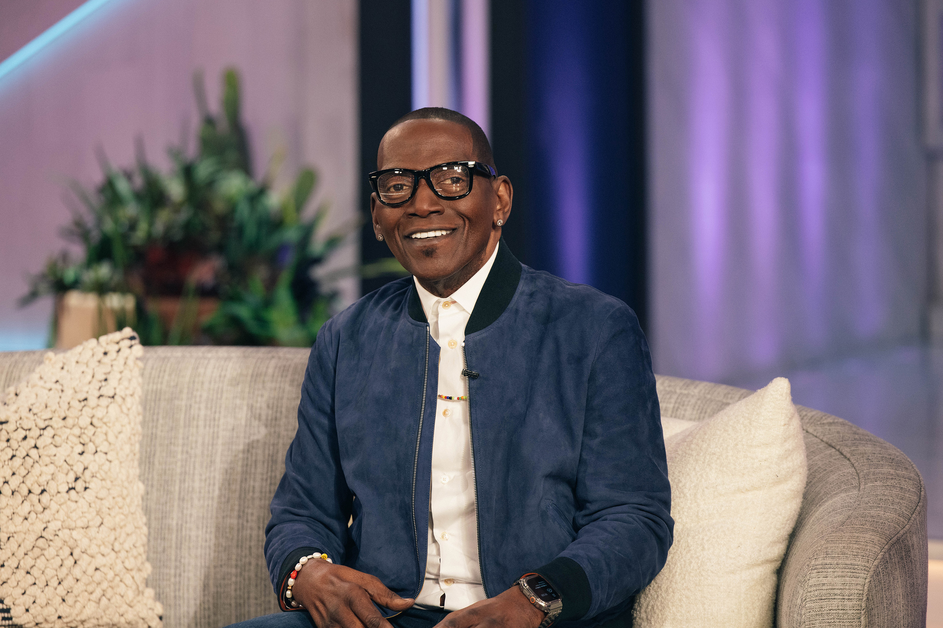 Seated on the set of “The Kelly Clarkson Show,” Randy Jackson smiles warmly in a blue suede-style jacket paired with dark-framed glasses.
