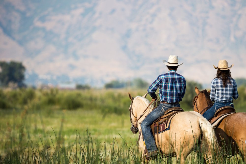 A man and woman horseback riding from behind overlooking wide open field and mountains. | Photo: Shutterstock.