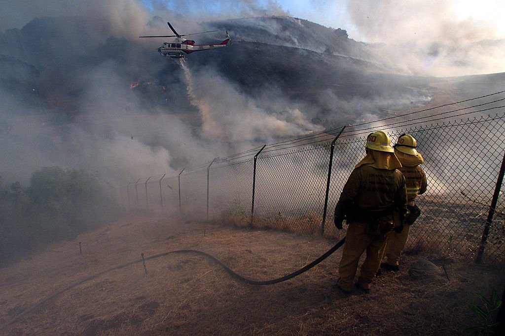 Die Feuerwehrleute von LA City beobachten, wie ein Hubschrauber auf einem brennenden Hügel in West Hills entlang der Zaunlinie zwischen LA und Ventura County einen Wassertropfen erzeugt. | Quelle: Getty Images