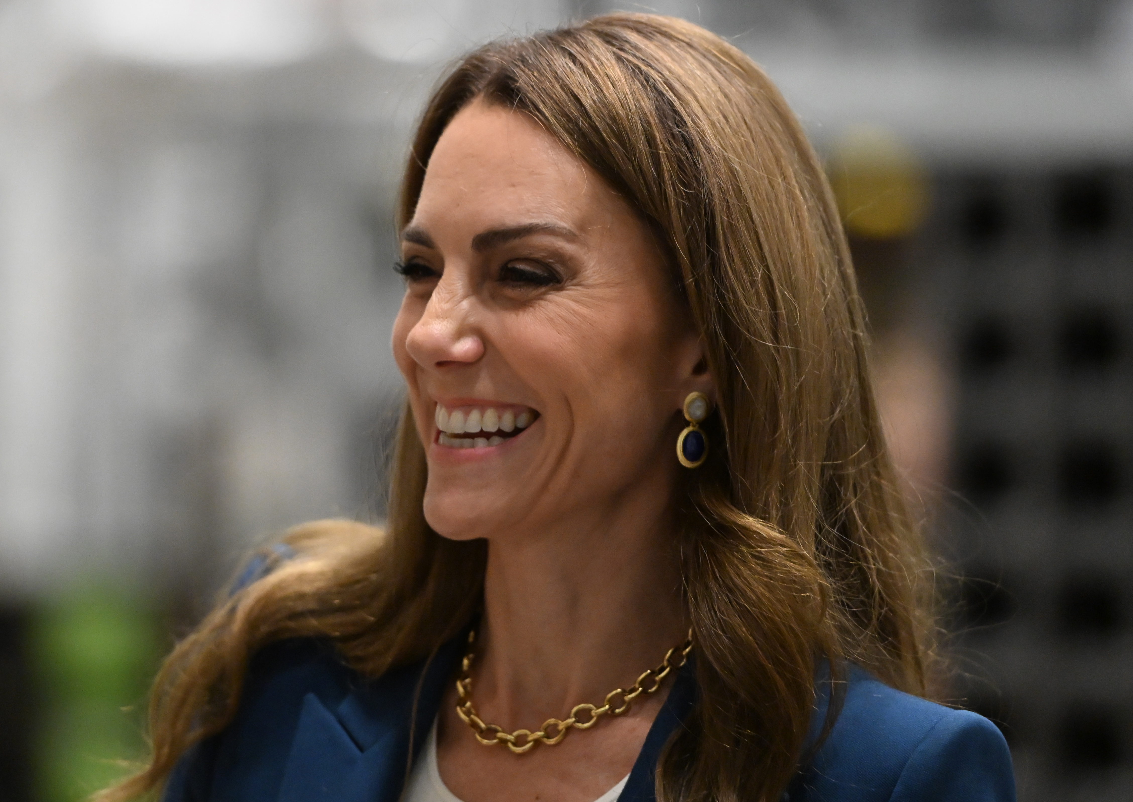 Princess Catherine smiles warmly during her museum visit on June 10, 2025, wearing a royal blue blazer, bold gold chain necklace, and blue drop earrings. Her soft waves and natural makeup complement the confident, polished tone of her appearance.