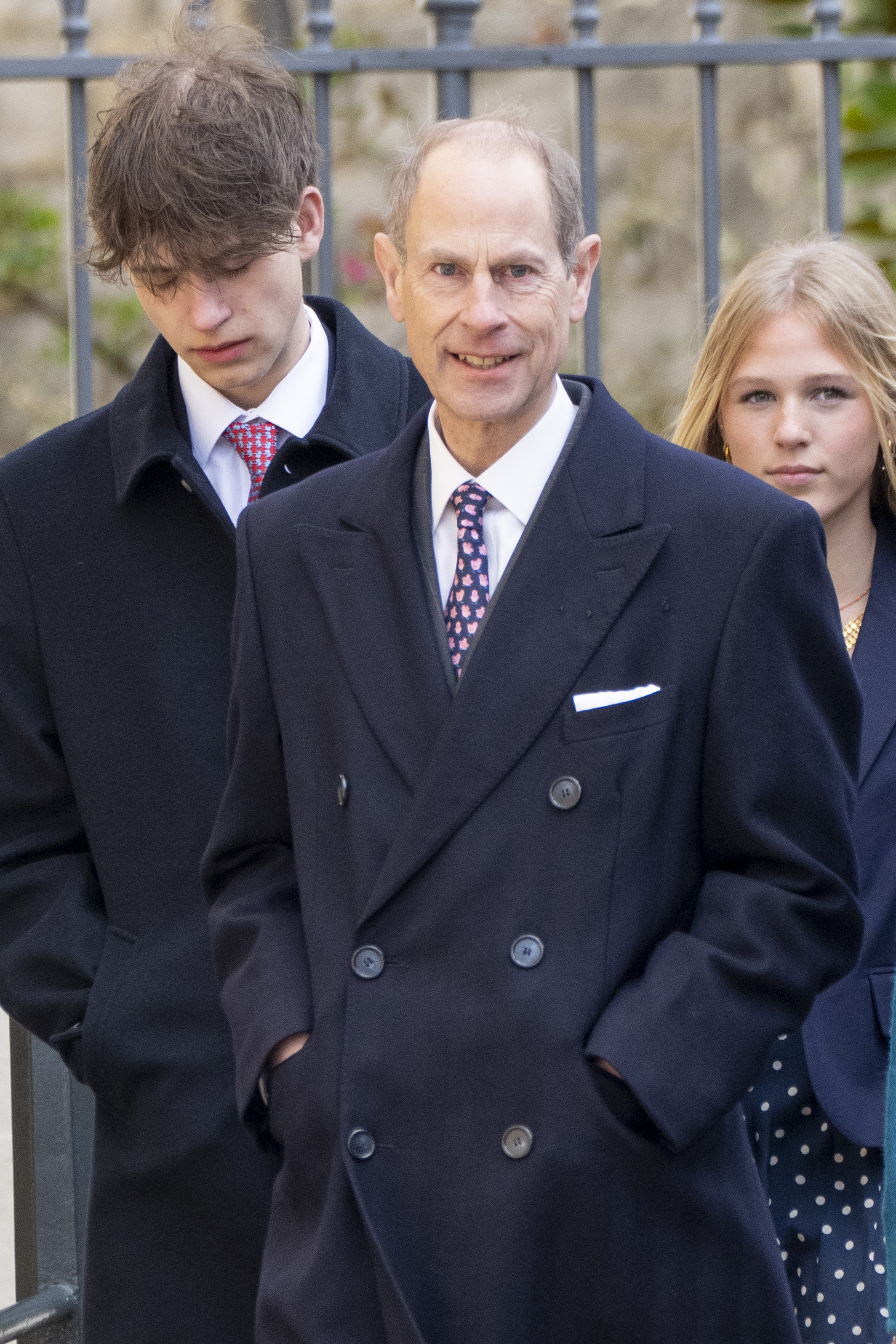 Prince Edward, Duke of Edinburgh, smiles as he makes his way through the Windsor Castle grounds on 5 April 2026, his son, James, Earl of Wessex, at his left shoulder. The teenager, dressed in a dark coat and a red patterned tie, walks with his hands in his pockets and his gaze cast downward — every inch the young royal finding his feet at a major family occasion.