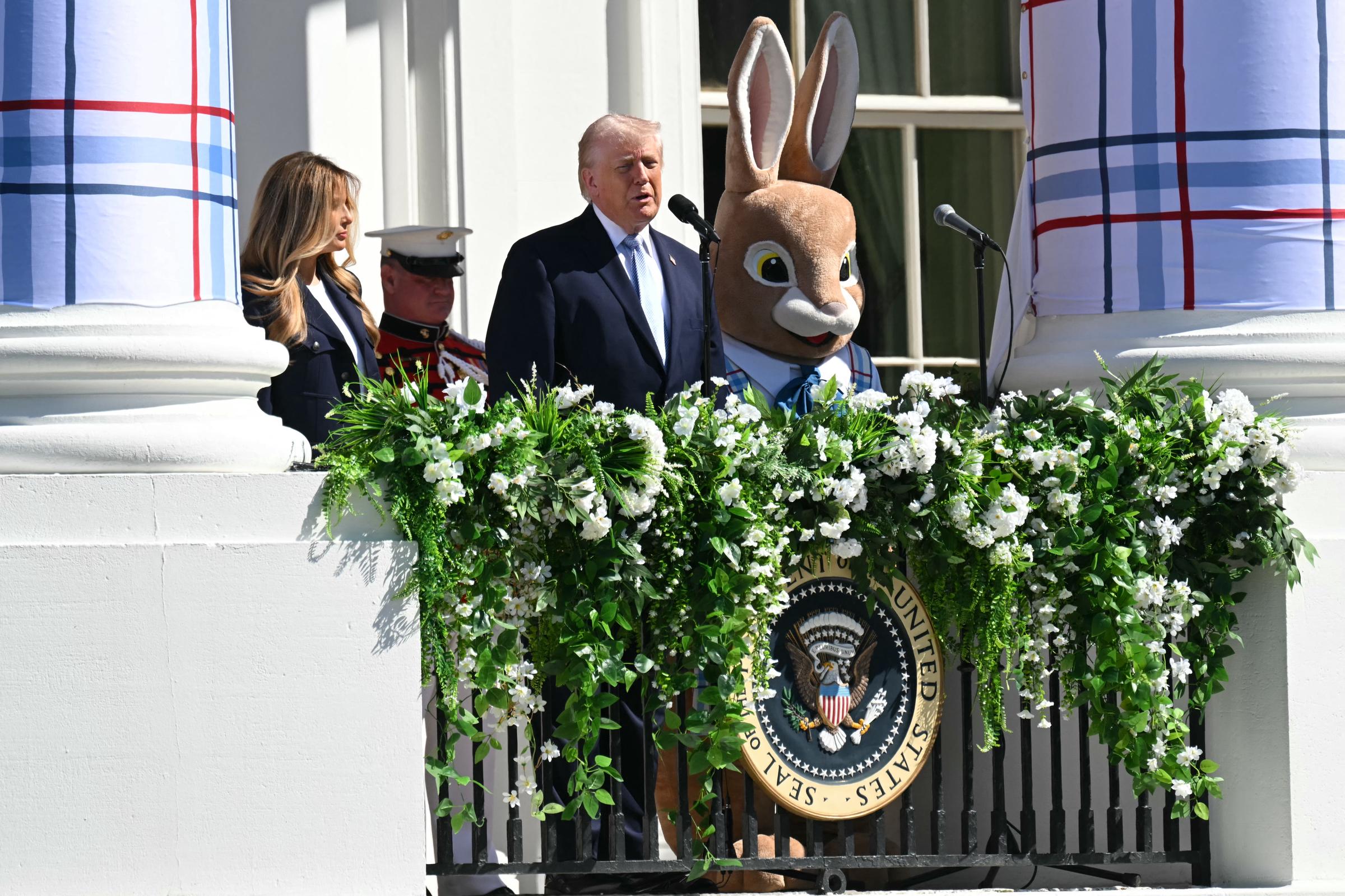 Donald Trump speaks from the White House balcony as Melania Trump and the Easter Bunny stand nearby, with greenery, plaid-wrapped columns, and spring flowers brightening the scene.