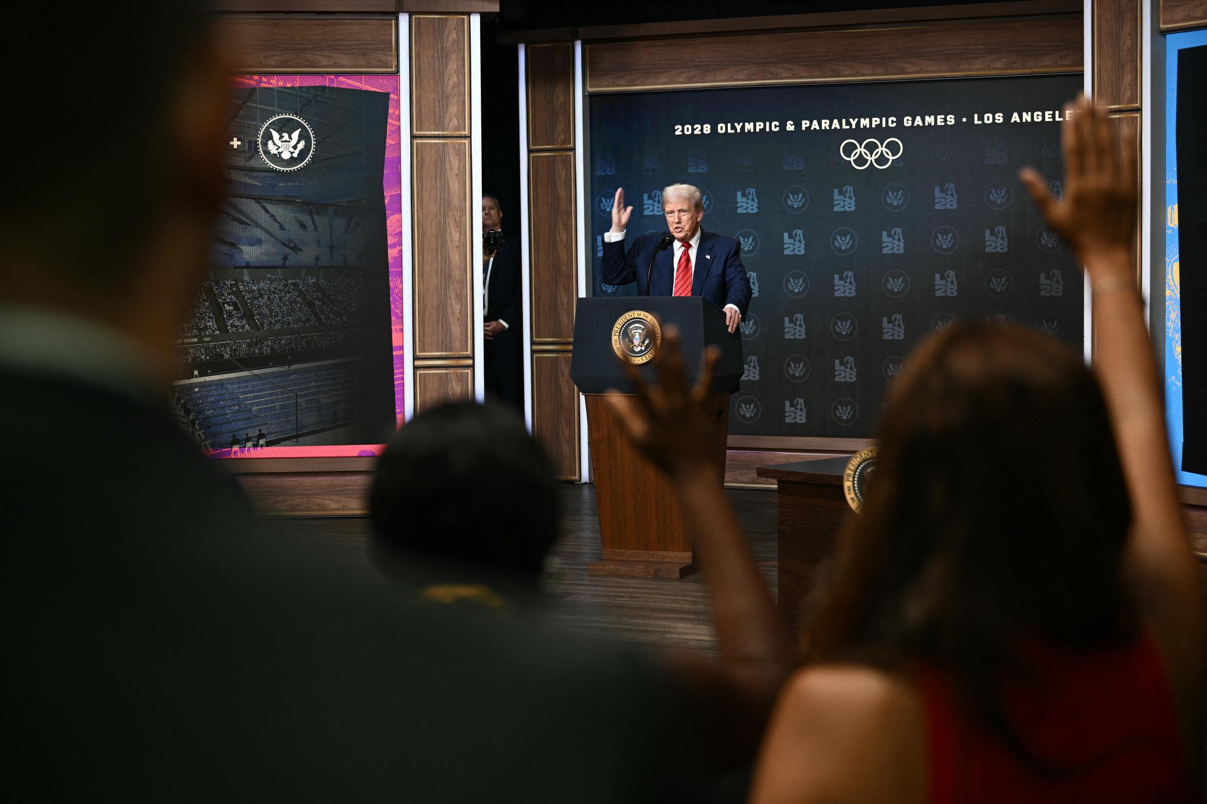 Following his rooftop walk at the White House, Donald Trump speaks from the South Court Auditorium podium, answering questions after signing an executive order to form a 2028 Olympics task force, while members of the press raise their hands.