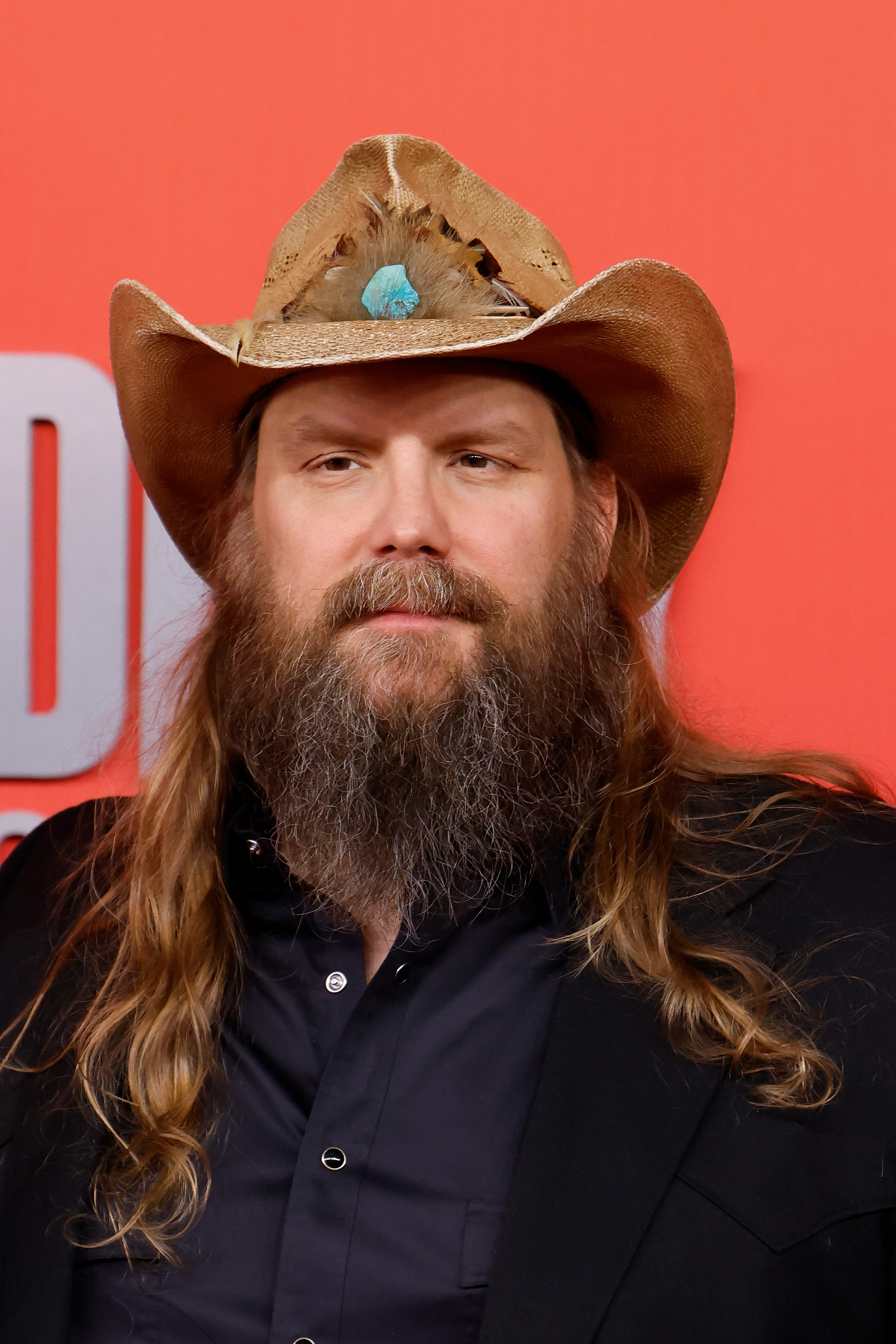 Chris Stapleton attends the 2025 ACM Awards at Omni Frisco Hotel in Frisco, Texas. He sports his signature long hair and beard, wearing a classic black button-up and a feather-accented cowboy hat for a rugged country look.