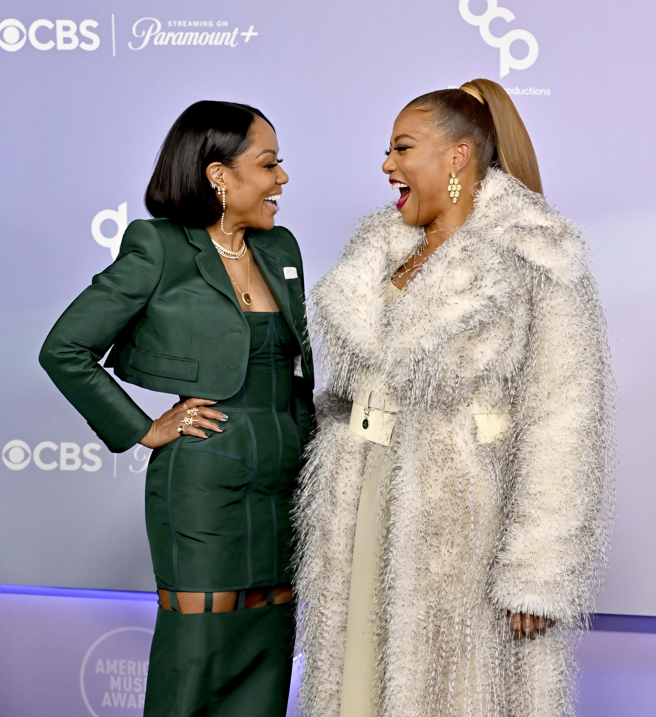 Eboni Nichols and Queen Latifah share a joyful laugh while facing one another during a candid red carpet moment in front of the American Music Awards media backdrop.