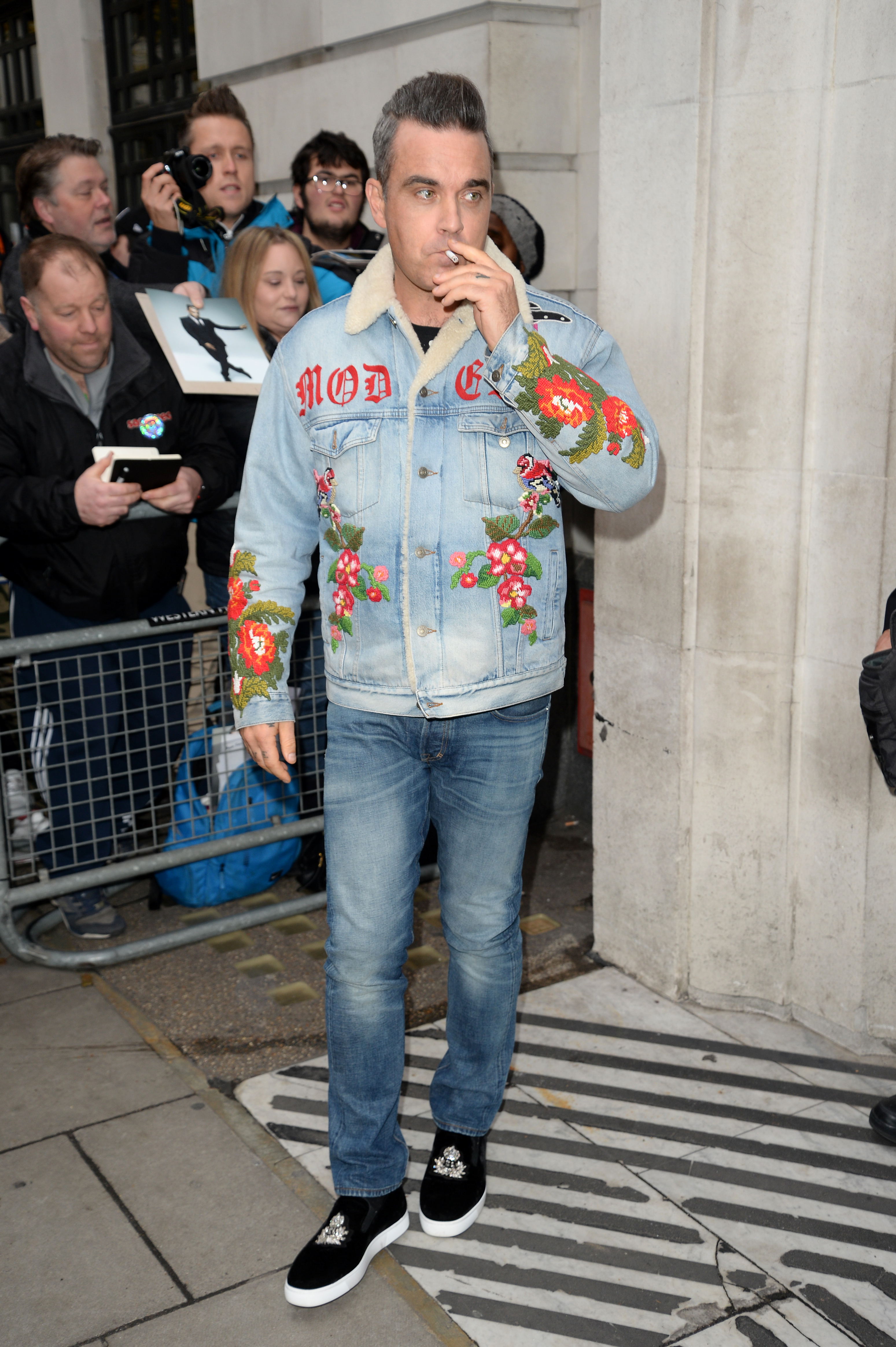 Robbie Williams was spotted outside BBC Radio 2 on December 8, 2017, in London, England. Wearing a floral denim jacket and smoking a cigarette, he made a bold style statement while greeting fans gathered outside the studio.
