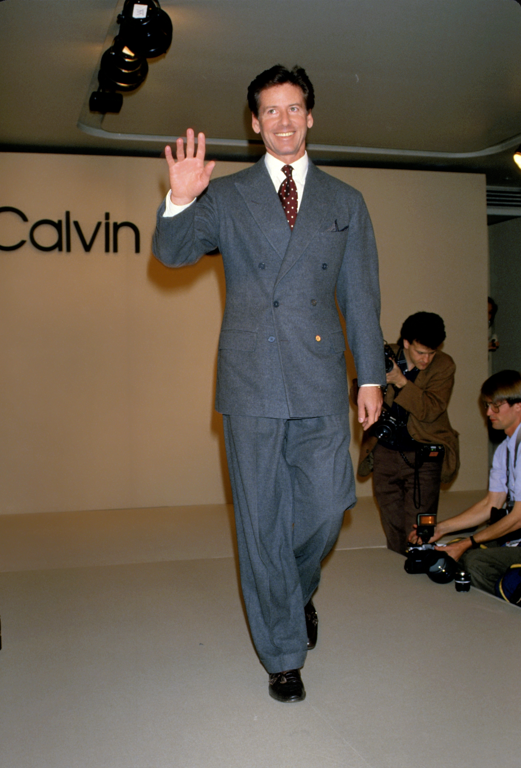 Beaming with confidence, Calvin Klein steps onto the runway during New York Fashion Week circa 1988, embodying the sharp tailoring and minimalist aesthetic that defined his brand. Dressed in a classic double-breasted suit, the designer greeted photographers and onlookers with a wave, marking another high point in a career that was already reshaping American fashion.