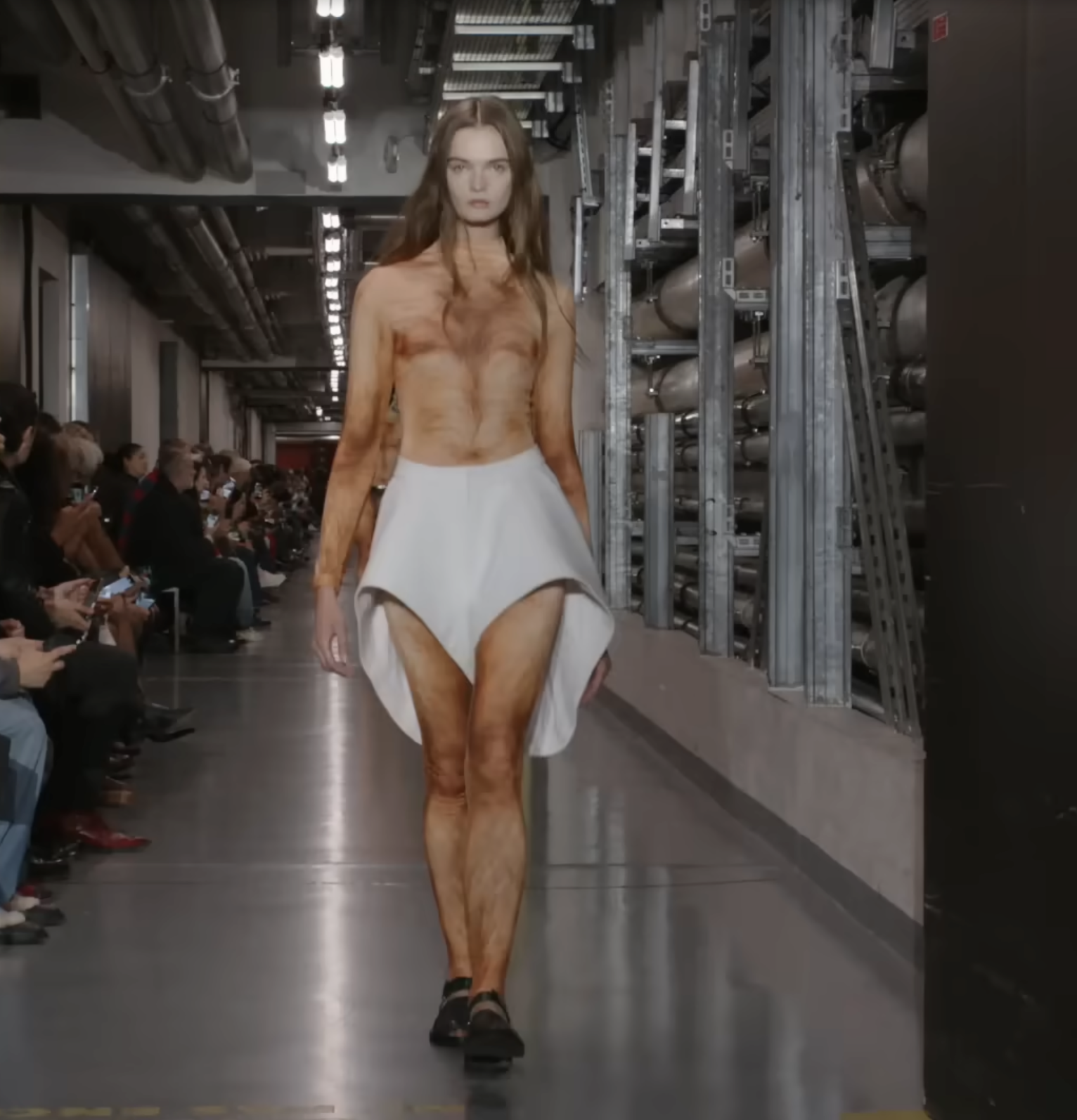 Another male model made a bold entrance in a striking white skirt with asymmetric high-and-low sections. It appears to be his only piece of clothing as he confidently strides down the runway. From a post dated October 5, 2025. Source: Youtube/@jeanpaulgaultier