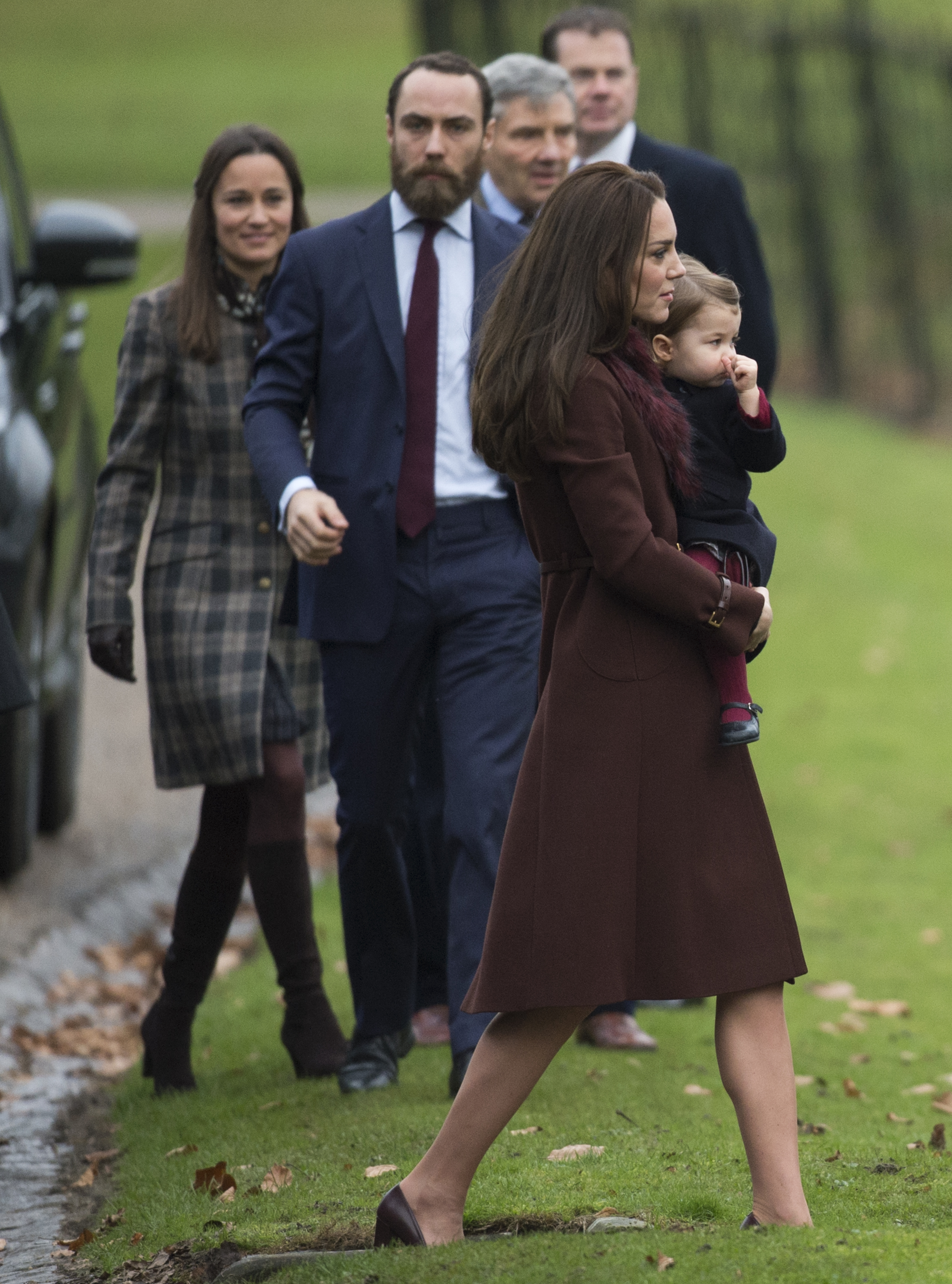 Princess Catherine walks to Christmas Day service in Bucklebury on December 25, 2016, carrying a young Princess Charlotte. She wears a deep burgundy coat and matching pumps, with her hair styled in a sleek brunette blowout. Her family walks behind her.