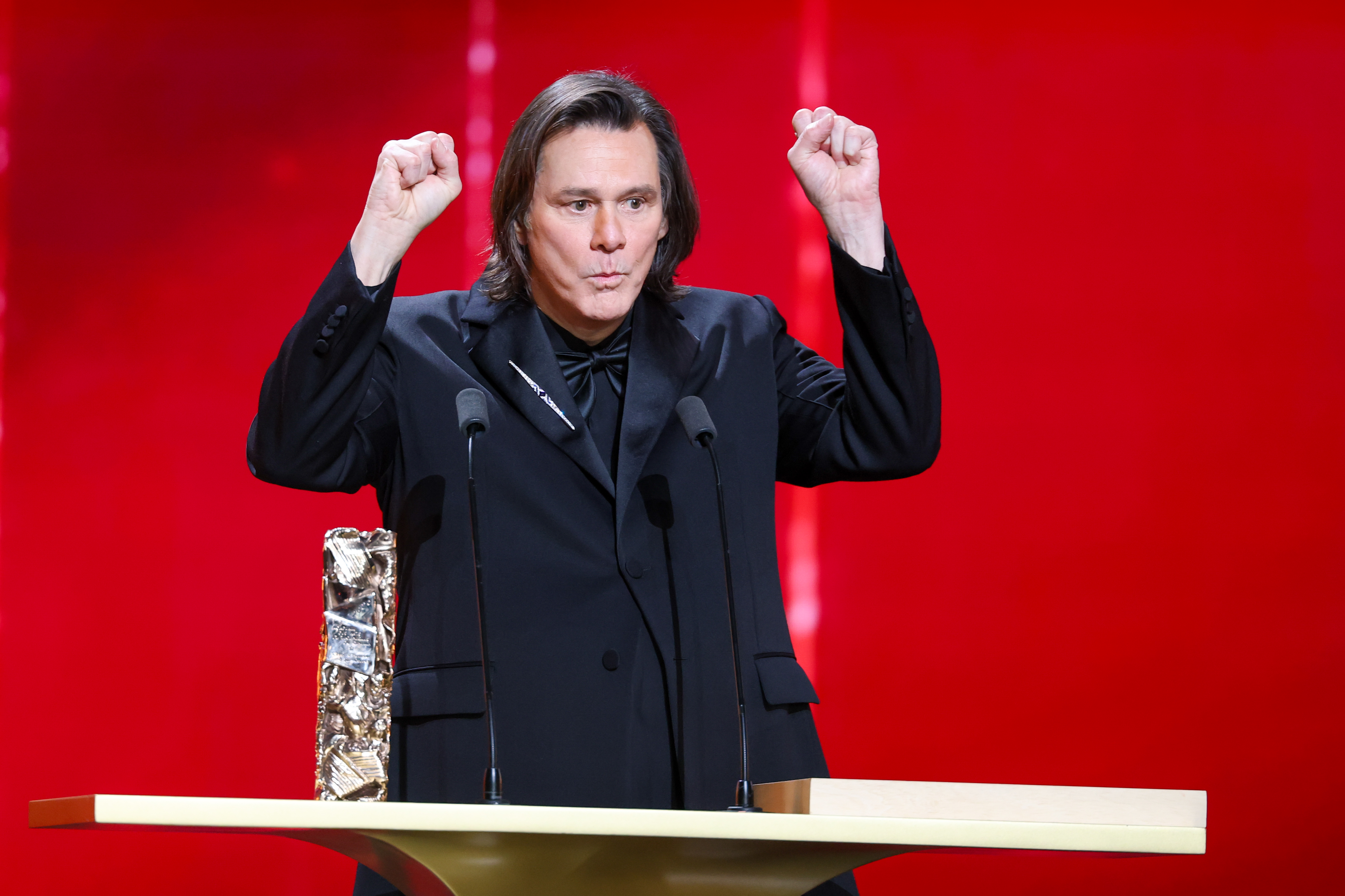 With both fists raised mid-gesture, Jim Carrey delivers an animated moment onstage. Dressed in a sharp black tuxedo, he stands behind the podium beside his Honorary César Award. The lively expression plays out during the 51st César Film Awards at L'Olympia in Paris, France.
