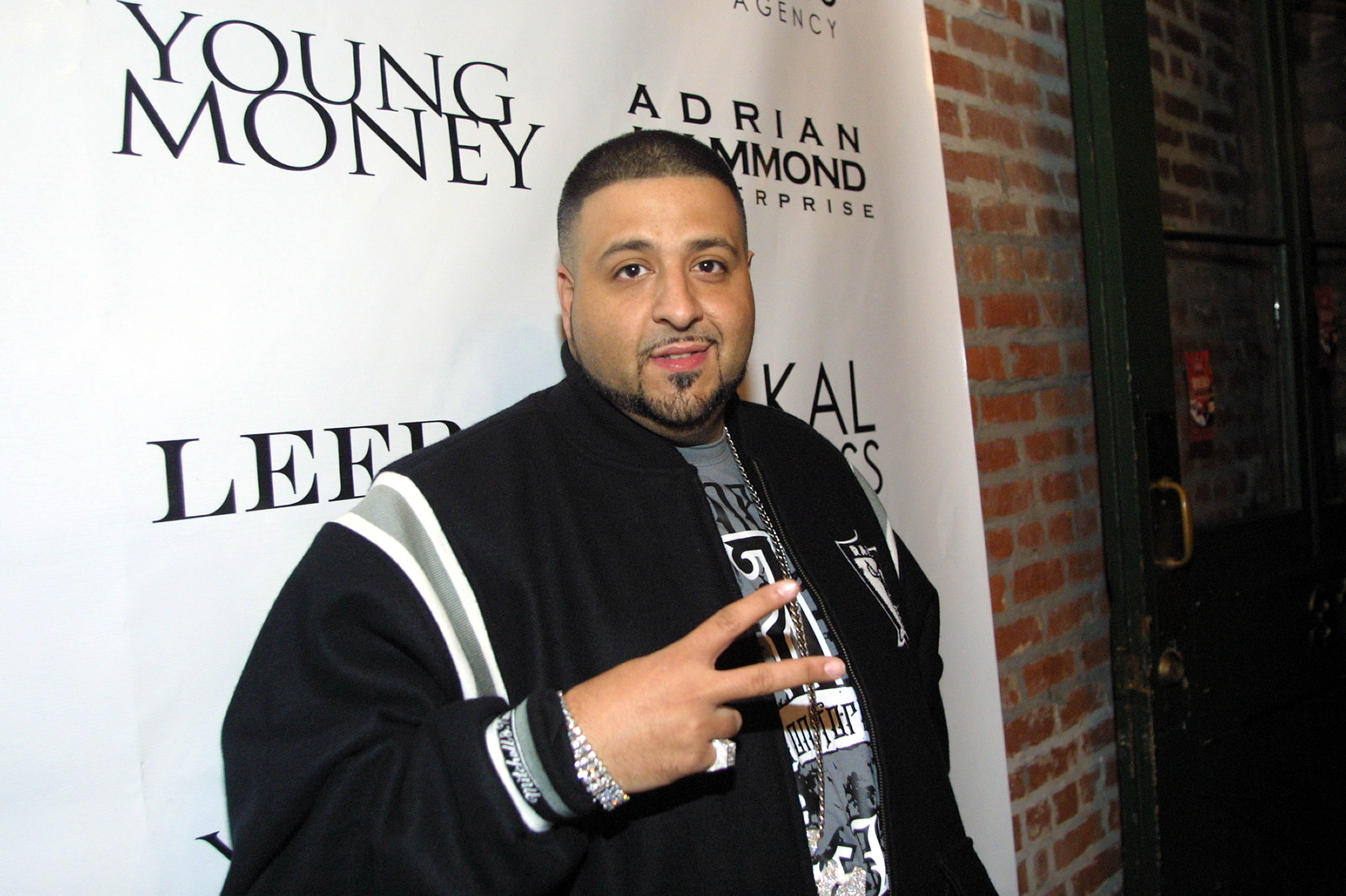 At a Young Money event, DJ Khaled poses in a black varsity jacket layered over a graphic tee, raising a peace sign toward the camera while silver accessories subtly catch the light.