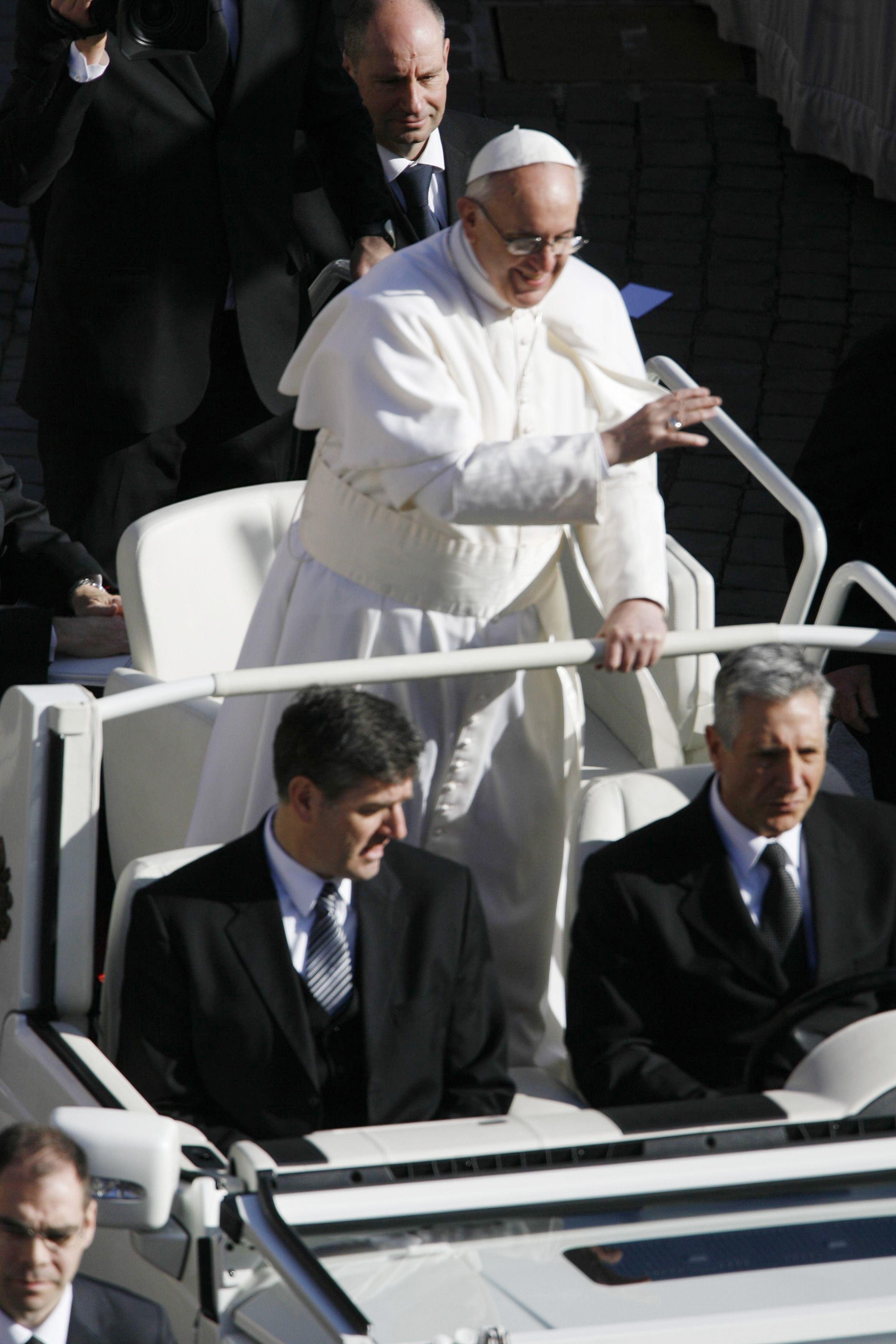 Pope Francis rides in the popemobile, smiling and waving in crisp white robes and skullcap, flanked by suited security detail amid a sea of dark coats