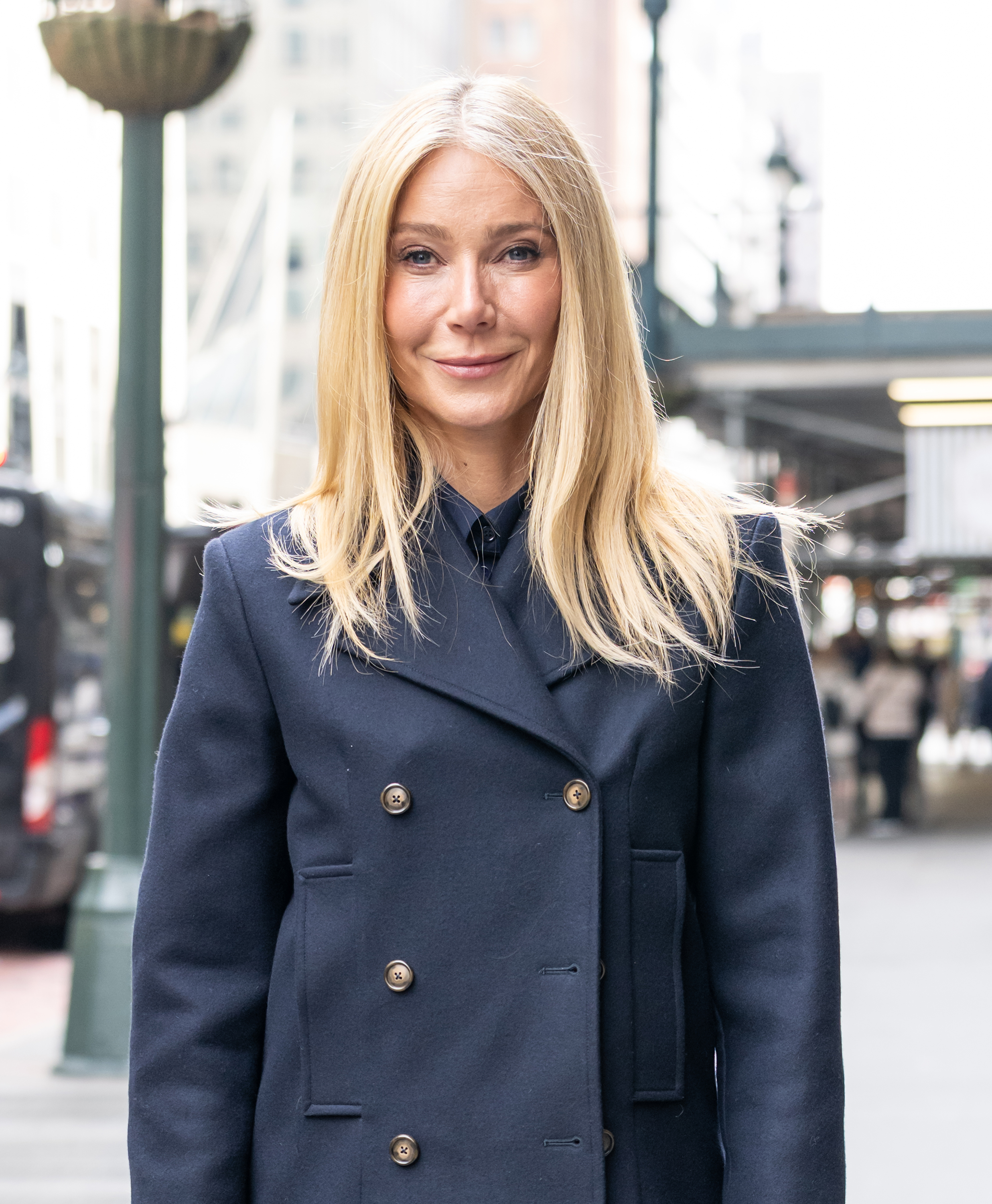 Gwyneth Paltrow is seen in a closer view, looking directly ahead with a subtle, controlled smile, her center-parted hair framing her face as the tailored navy look remains crisp.