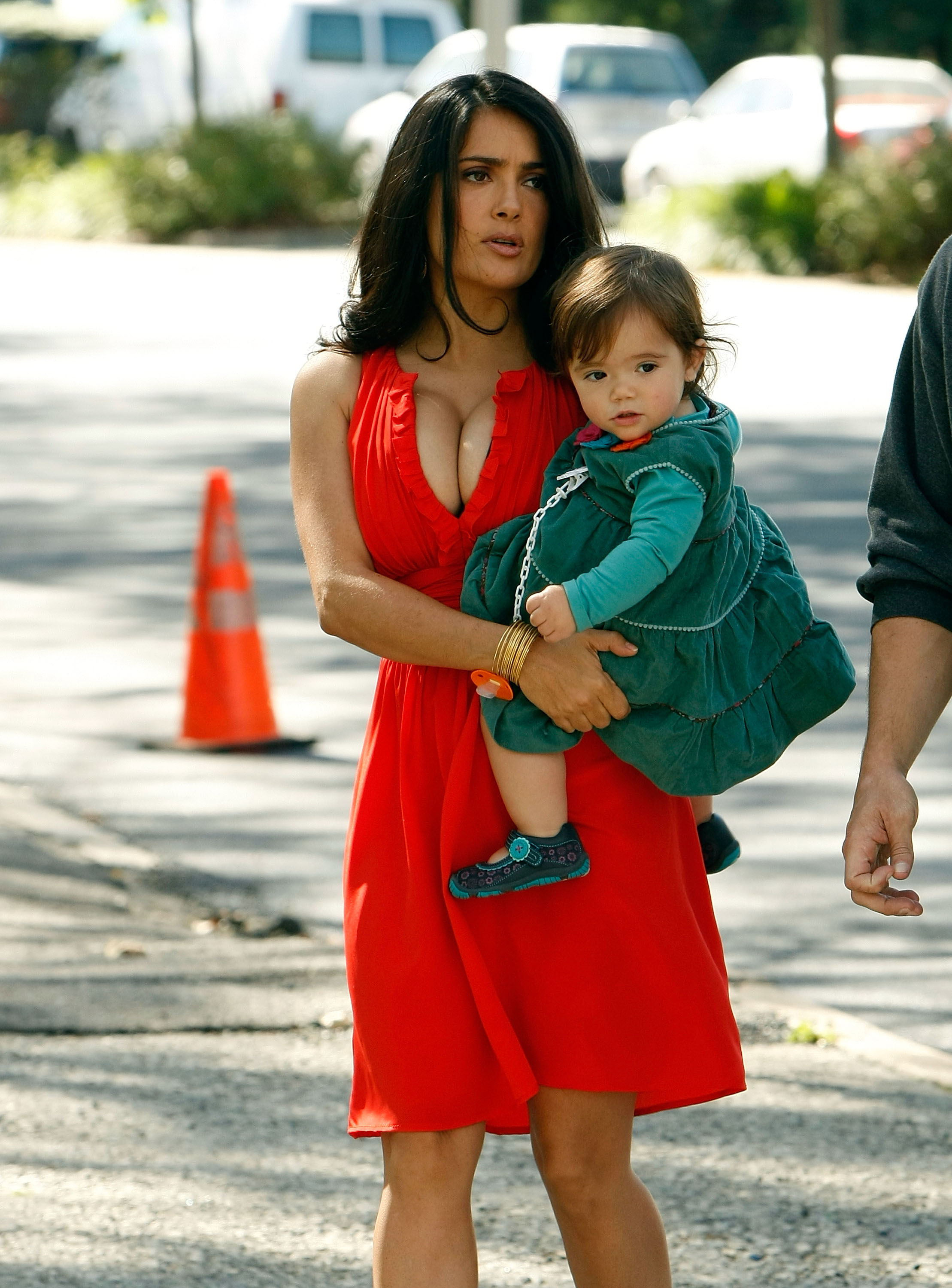 Salma Hayek is seen cradling baby Valentina Pinault in her arms while on the set of "30 Rock" in New York City. Wearing a bold red dress with gold accessories, Salma walks briskly across the pavement with a serious expression. Valentina, dressed in an emerald green tiered outfit with matching leggings and ballet flats, gazes calmly into the distance.