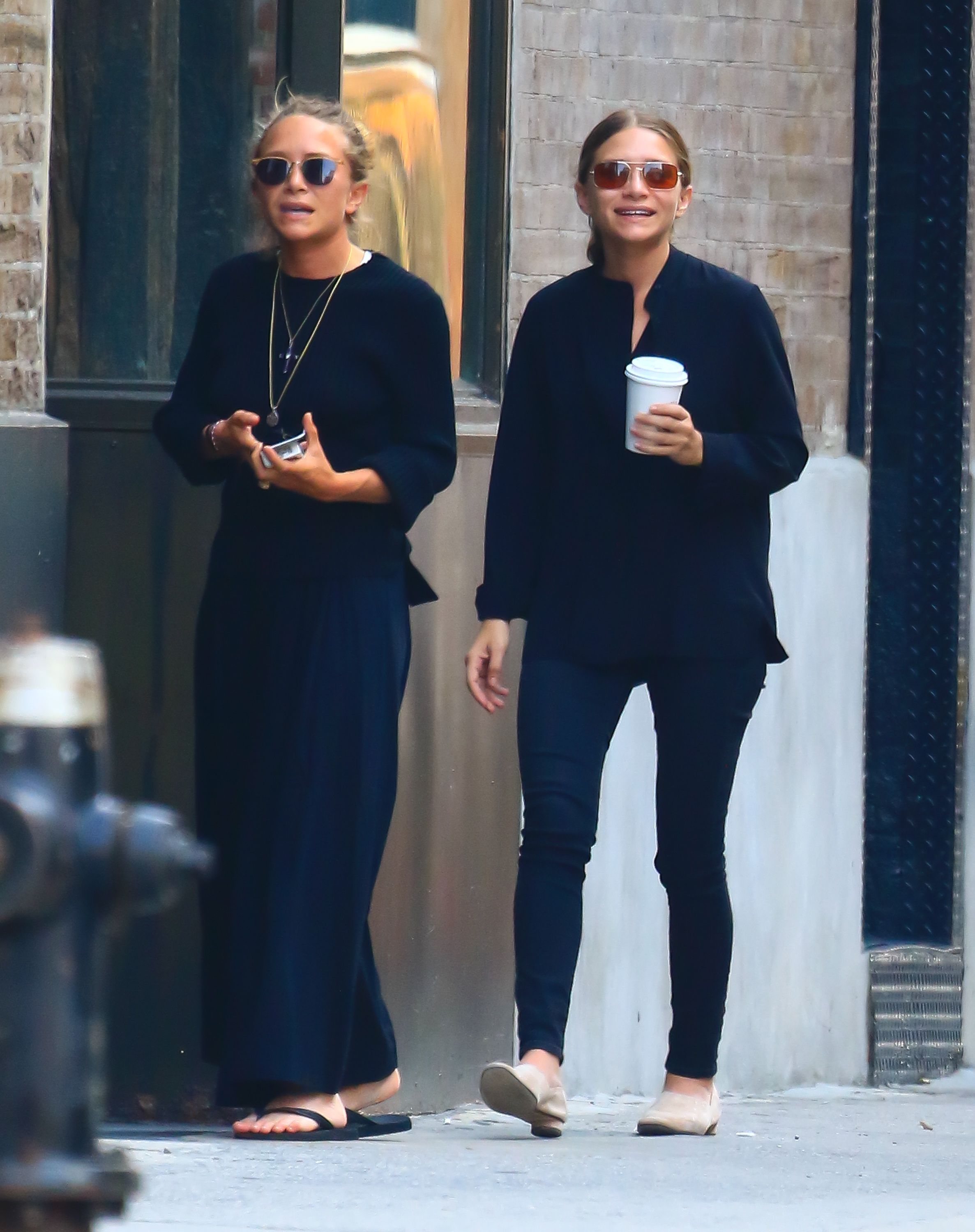 Mary-Kate Olsen and Ashley Olsen walk side by side in the city, both in dark, relaxed outfits and sunglasses. One holds a drink as they move casually down the street.