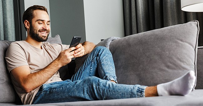 A man is texting on his phone, while lounging on the couch. | Photo: Shutterstock