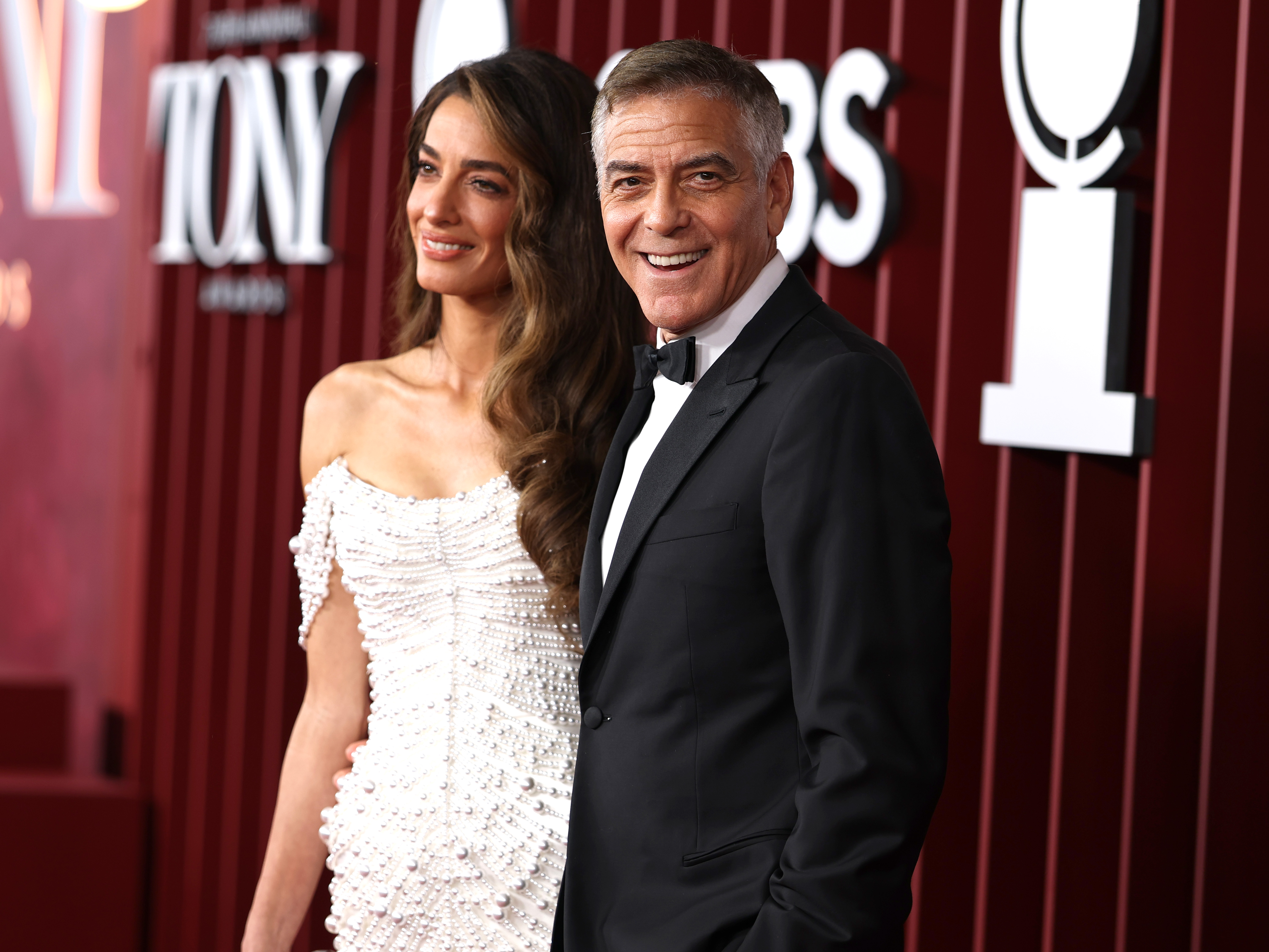 George Clooney smiles softly for the cameras while Amal Clooney stands beside him. Her pearl-draped gown glistens under the lights as they pose together on the Tony Awards red carpet.