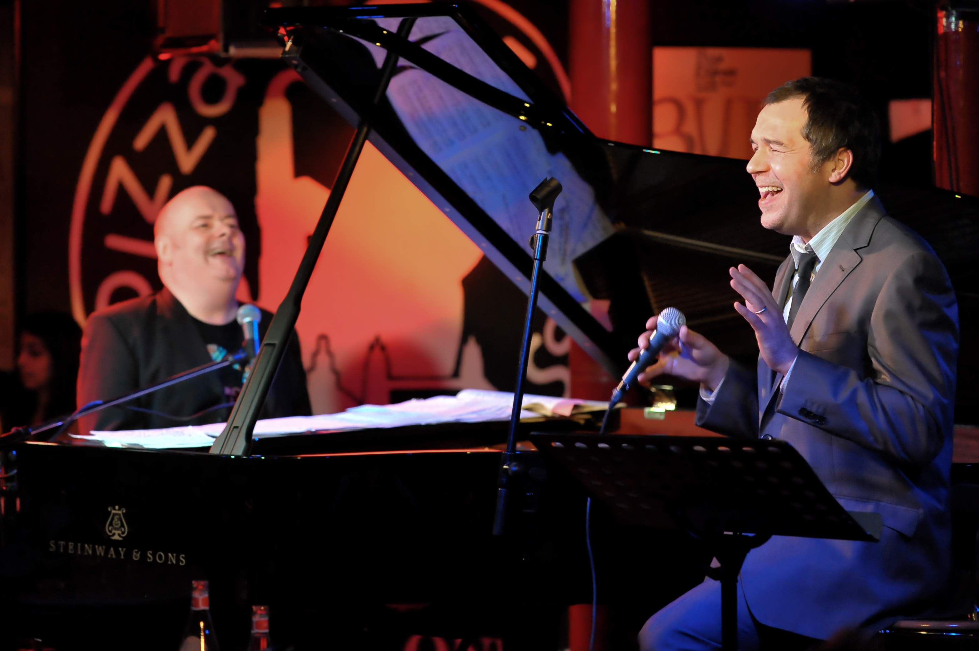 Brookstein shares the spotlight with his pianist at the same Pizza Express Jazz Club set, the pair caught mid-song beside a gleaming Steinway & Sons grand piano, the warm amber glow of the intimate venue surrounding them.