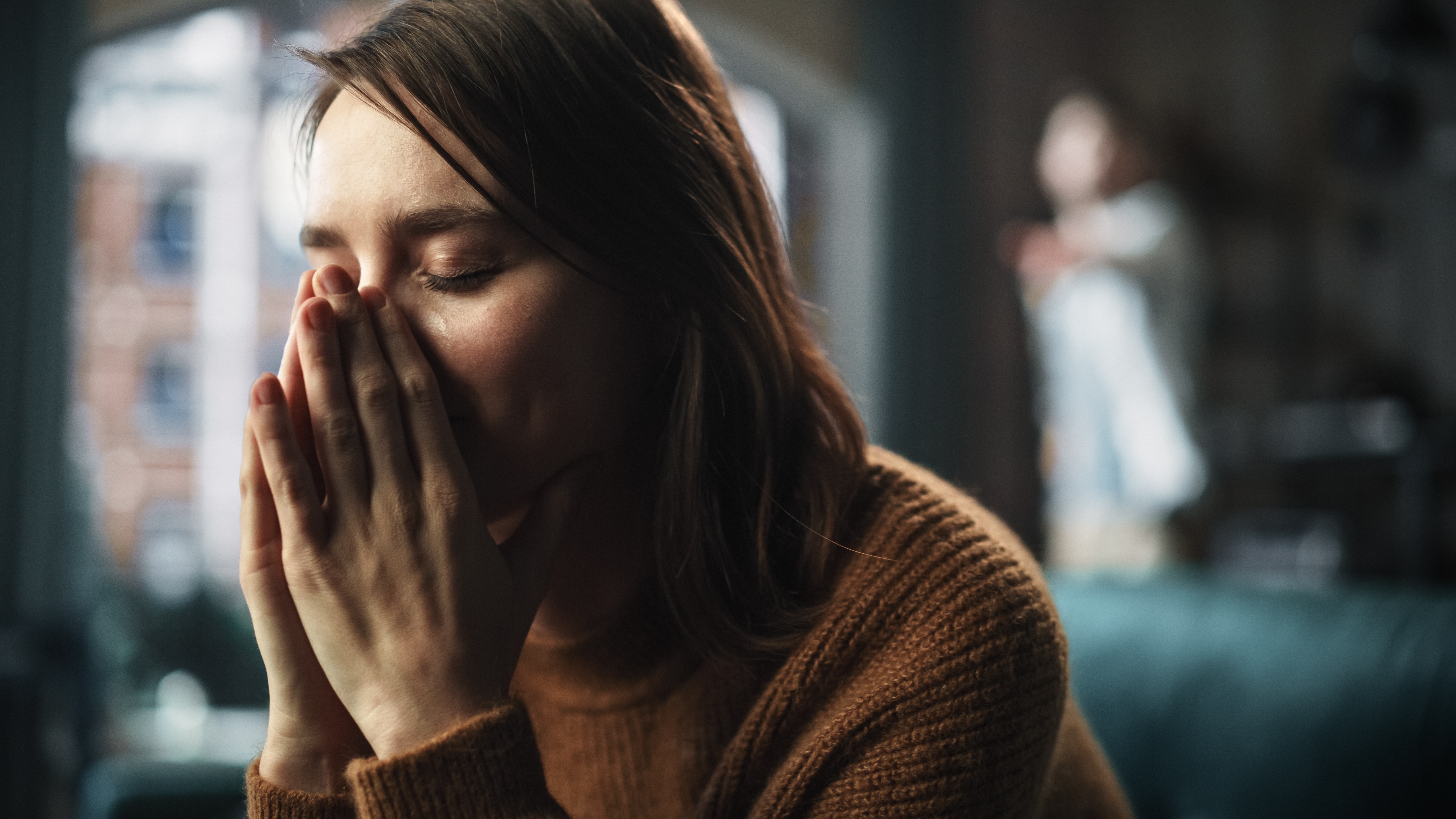 An upset woman covering her face while sitting on a couch with someone standing in the background | Source: Shutterstock