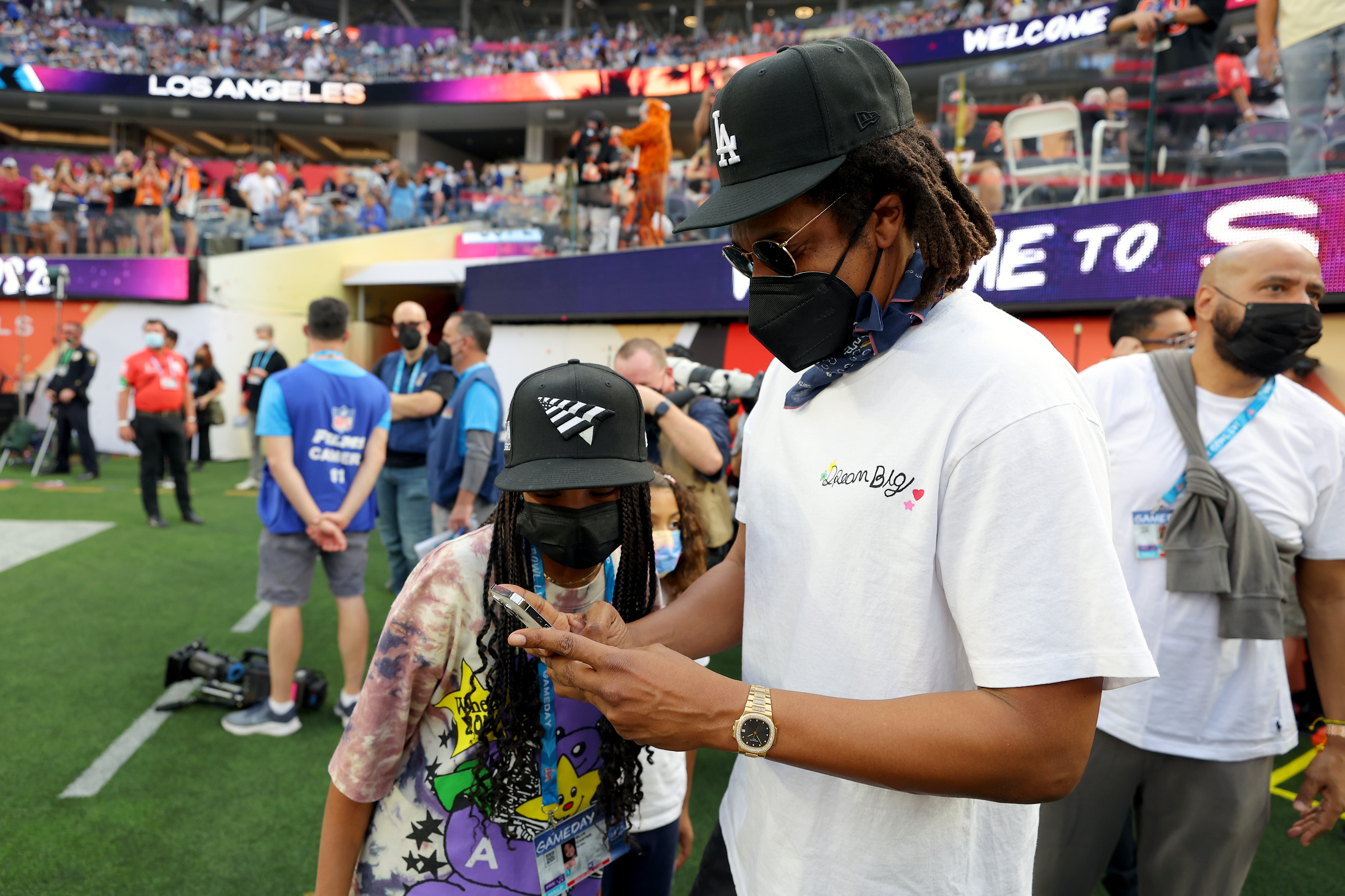 Jay-Z pauses on the sideline to show Blue Ivy something on his phone, drawing her attention amid the pregame activity. The interaction offers a quiet, candid father-daughter exchange.