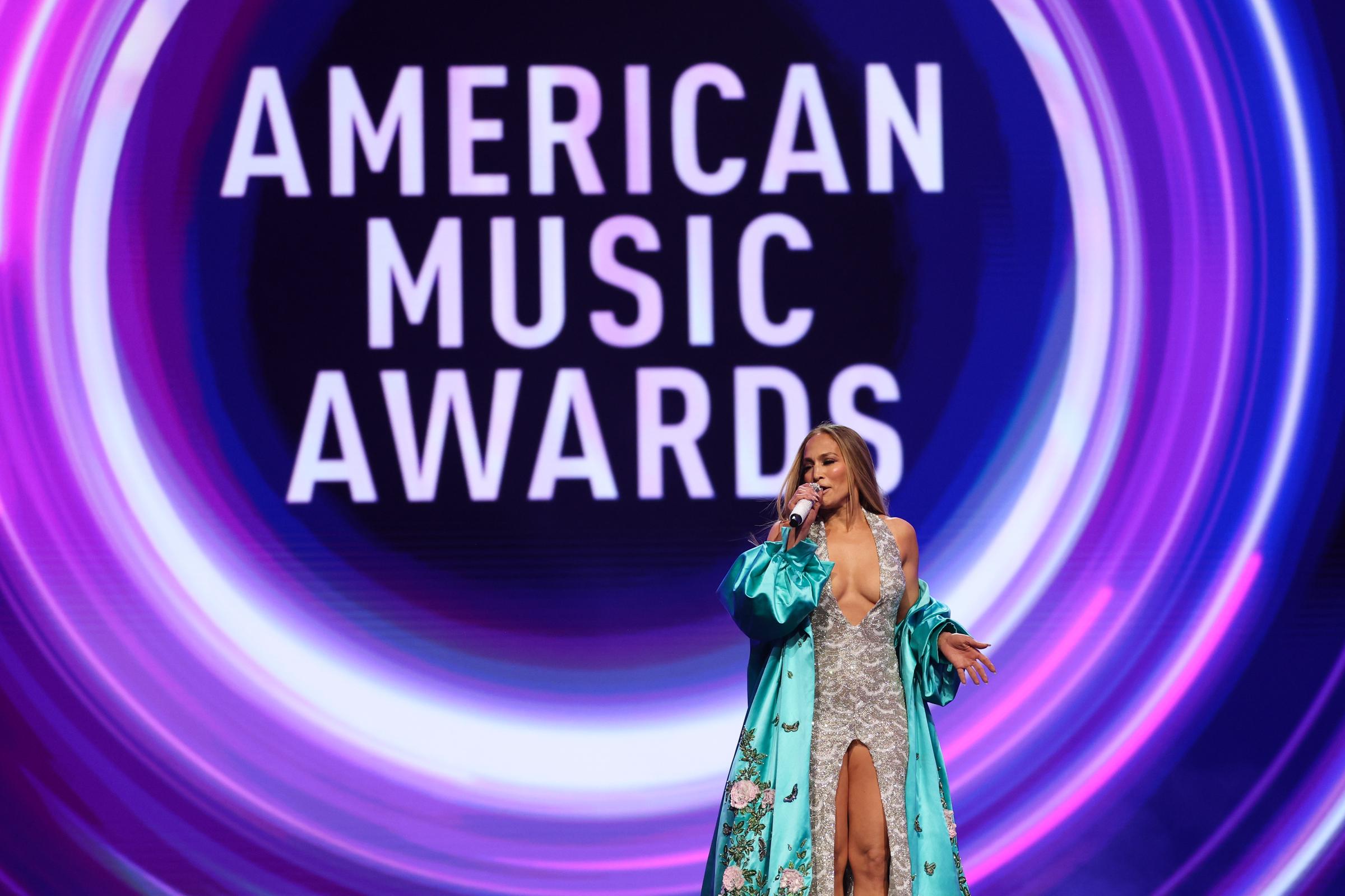 Mid-presentation, Jennifer Lopez raises one hand while speaking, the glitter of her gown and flow of her embroidered robe standing out against the bold, swirling AMAs stage graphics.