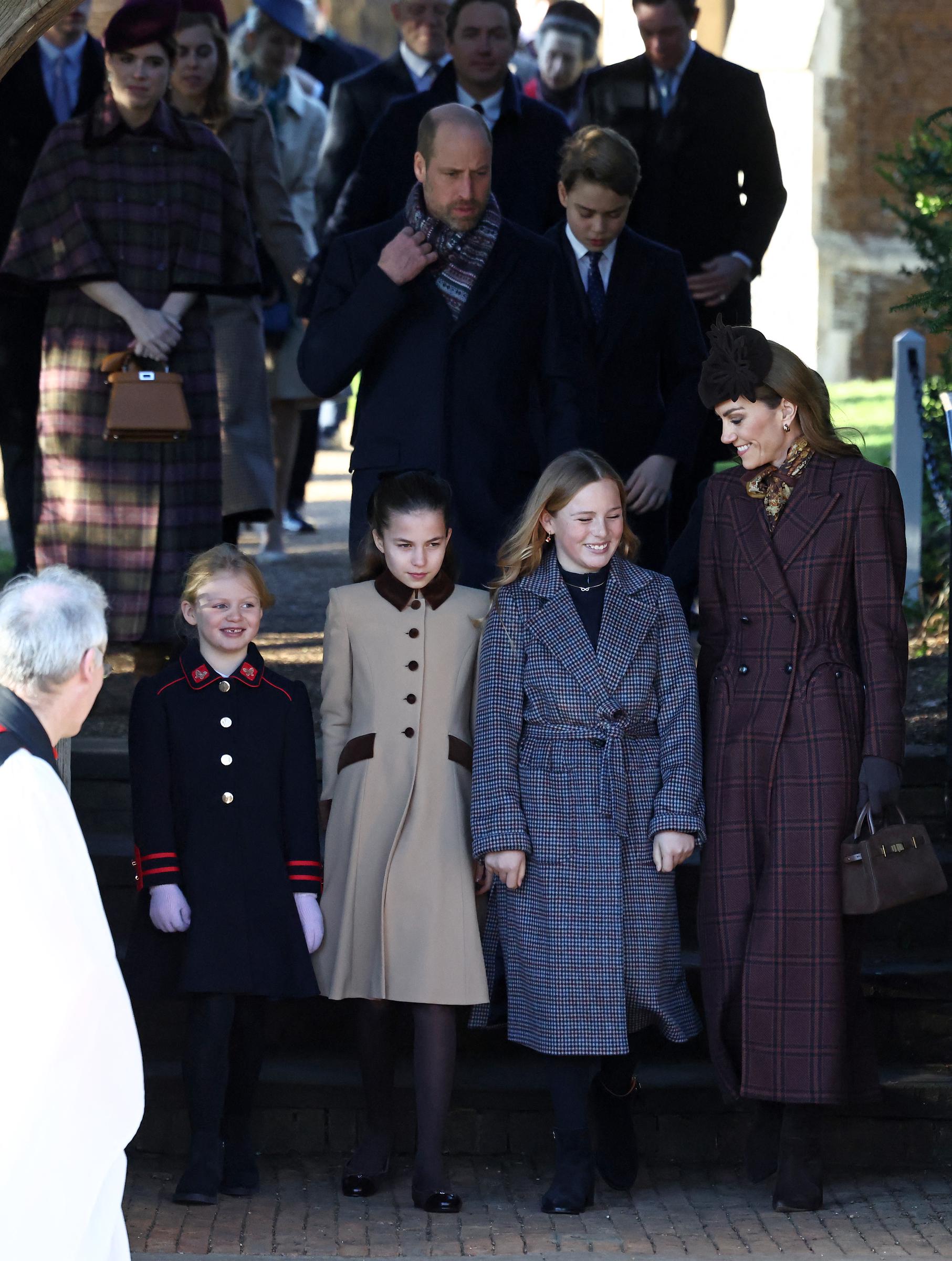 Catherine, Princess of Wales, smiles as she walks alongside children of the Royal Family, including Princess Charlotte, after the Christmas Day church service at Sandringham. Behind them, William, Prince of Wales, is seen adjusting his scarf — a gesture that body language experts later highlighted amid claims of social distance during the family gathering.