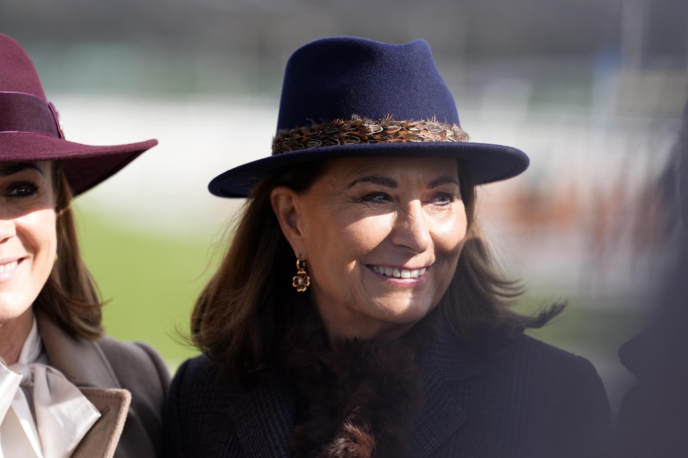 Beaming in the Gloucestershire sunshine, Carole Middleton sports the feather-trimmed Hicks & Brown blue fedora — the very hat last seen on the head of the Princess of Wales at Sandringham in 2020 — its distinctive pheasant-feather band catching the light as her floral drop earrings frame a radiant, rosy-lipped smile at Cheltenham Racecourse on 11 March 2026.