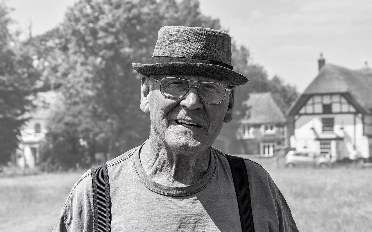 A black-and-white image of an old man wearing spectacles and a hat while standing by himself | Photo: Pixabay/shauking