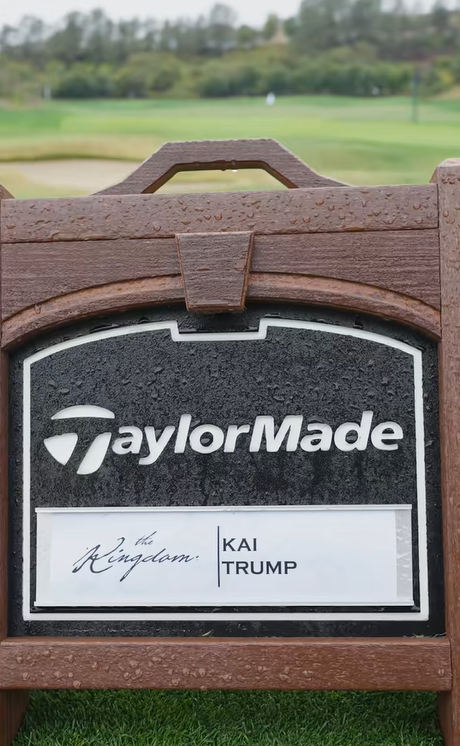 A TaylorMade sign featuring Kai Trump's name stands against a blurred fairway, dotted with raindrops on its wooden frame | Source: Tiktok/thekaitrumpgolfer