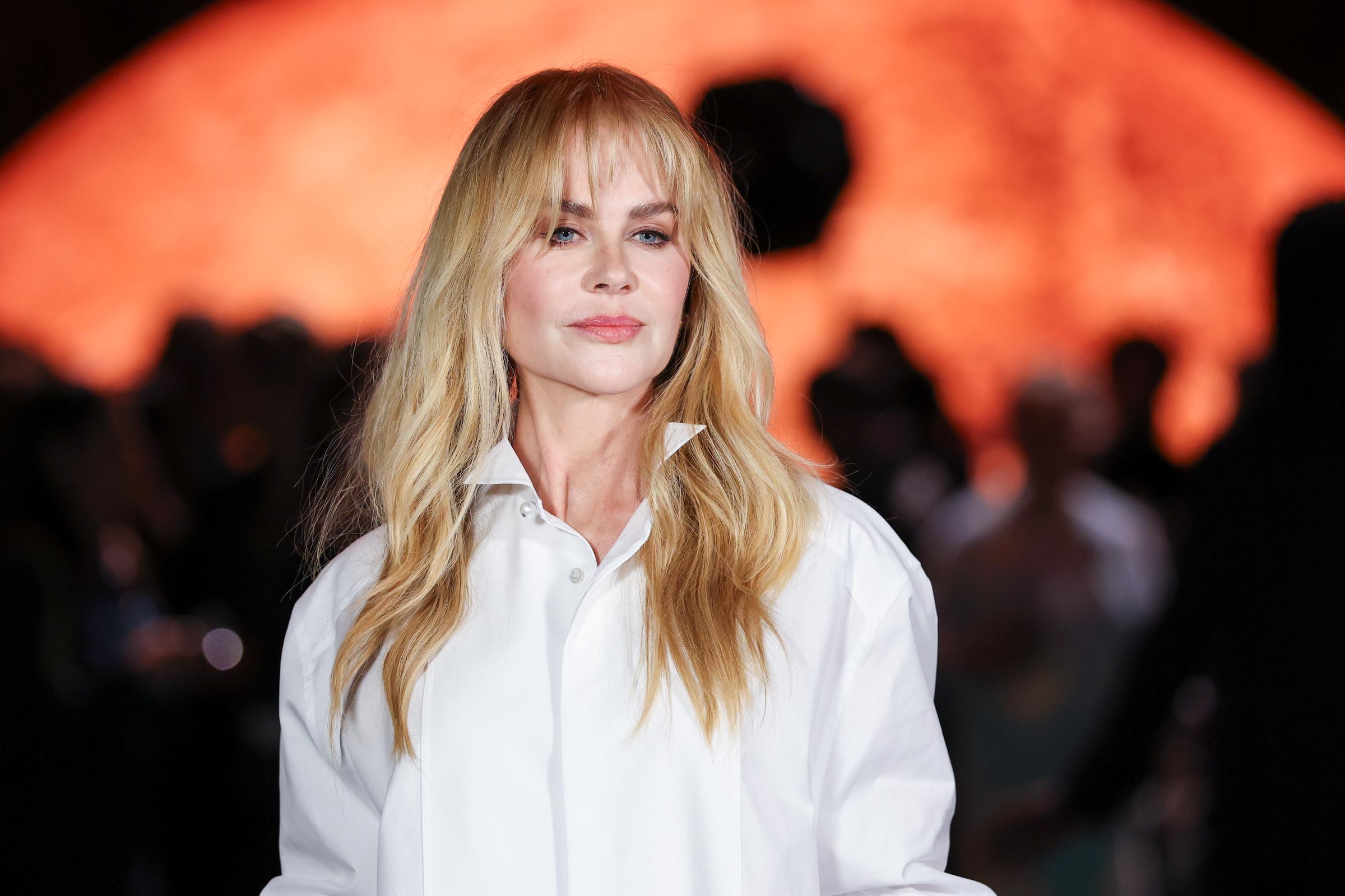 Nicole Kidman gazes into the distance as she debuts her bangs. The orange-hued background adds intensity to the soft look — her long blonde locks and sharp collar framing her face with sleek sophistication.