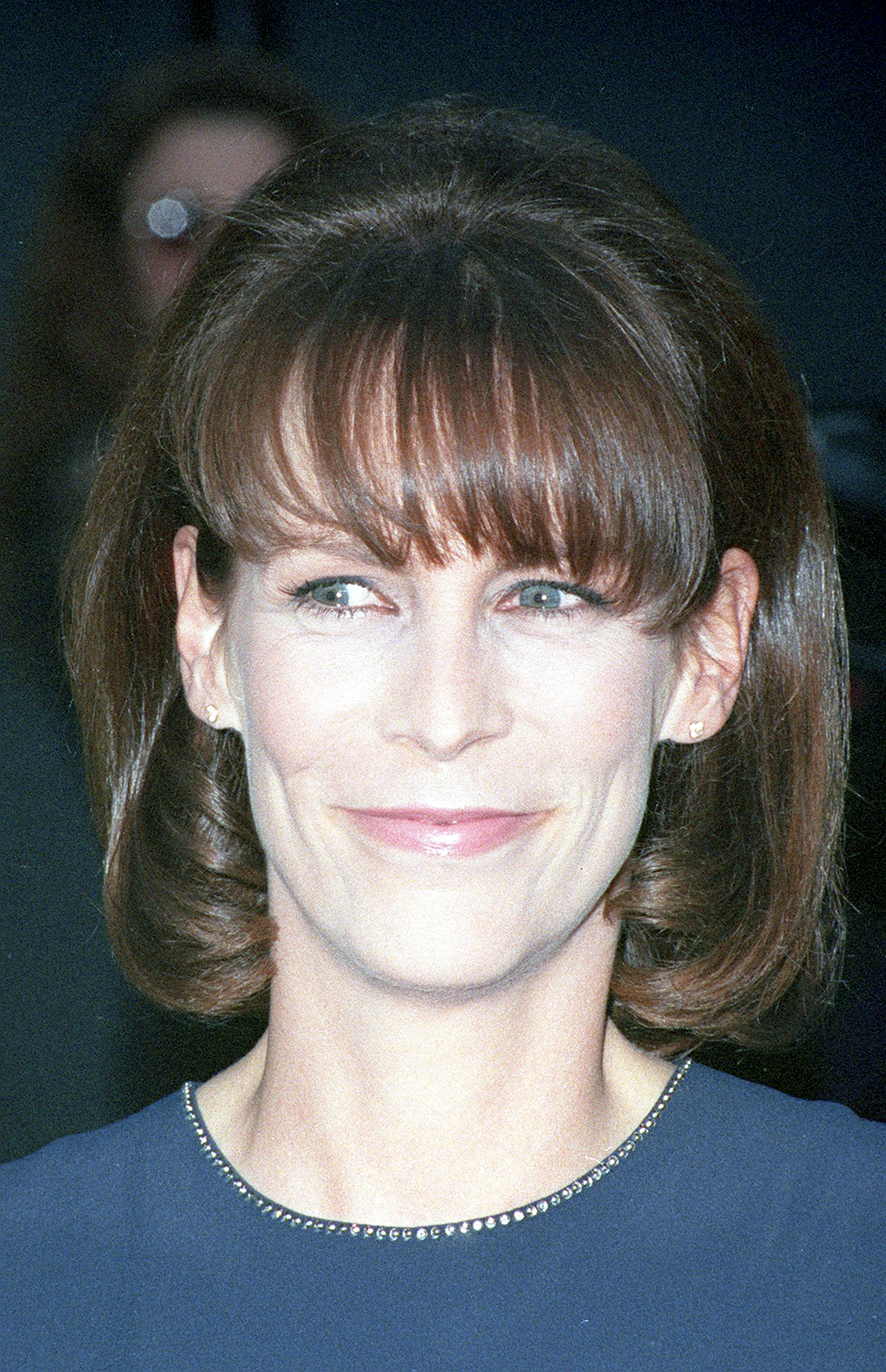 Jamie Lee Curtis showcases a polished mid-'90s look with voluminous shoulder-length hair and soft bangs, paired with a navy dress edged in subtle sparkle.