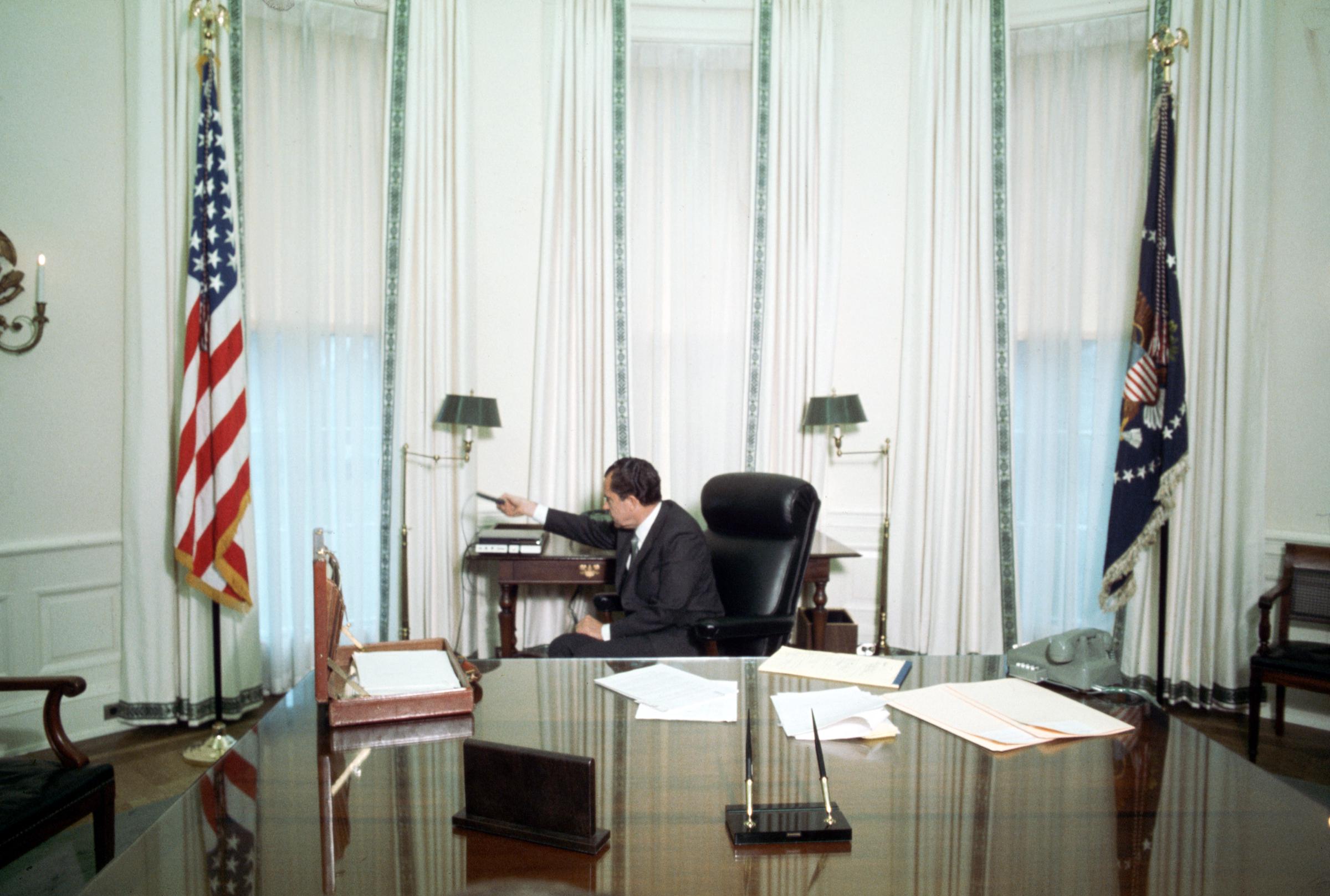 On January 21, 1969, President Richard Nixon can be seen demonstrating a tape recorder in a sparsely decorated Oval Office, marked by white drapes, twin flags, and a nearly bare desktop.