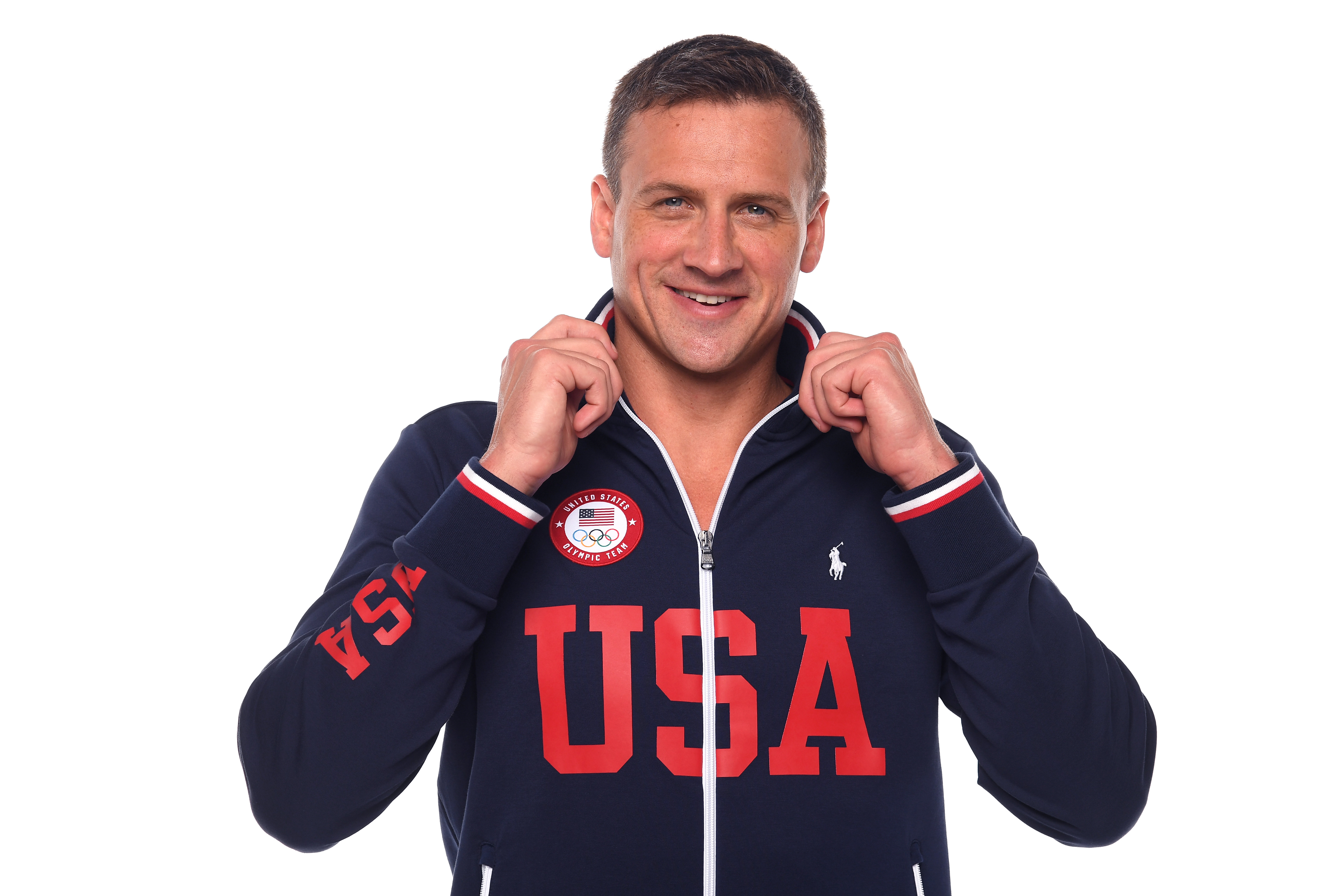 Ryan Lochte poses during a Team USA Olympic portrait shoot while wearing a navy zip-up jacket emblazoned with bold red "USA" lettering. The swimmer grips the collar of the jacket and smiles confidently against a clean studio backdrop.