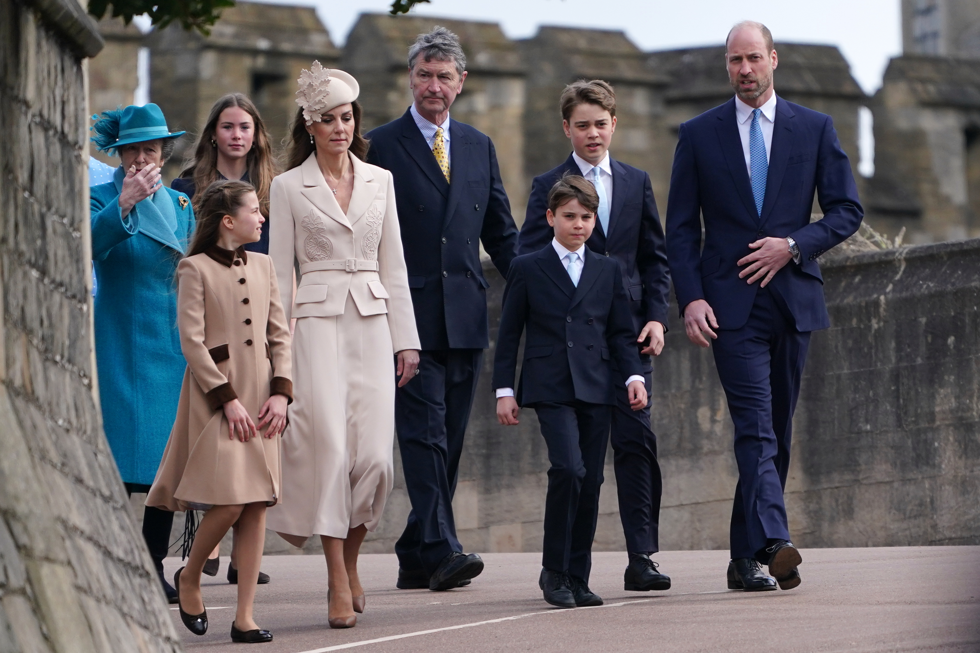 The Wales family makes its way along the Windsor Castle walkway, with Princess Charlotte glancing in the direction of the Princess of Wales. William, Prince of Wales, strides alongside Prince Louis and Prince George, while Sir Timothy Laurence walks behind the group and Anne, the Princess Royal, is visible in her teal coat on the far left.