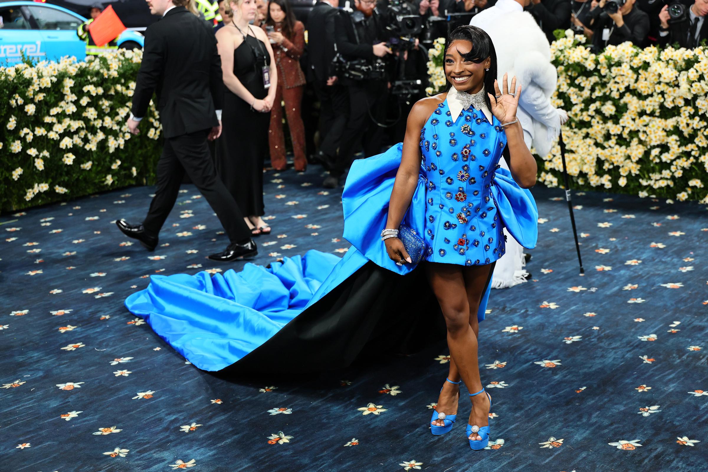 Simone Biles waves at the crowd while smiling, standing confidently in the center of the carpet. Her long train gathers elegantly at her feet.