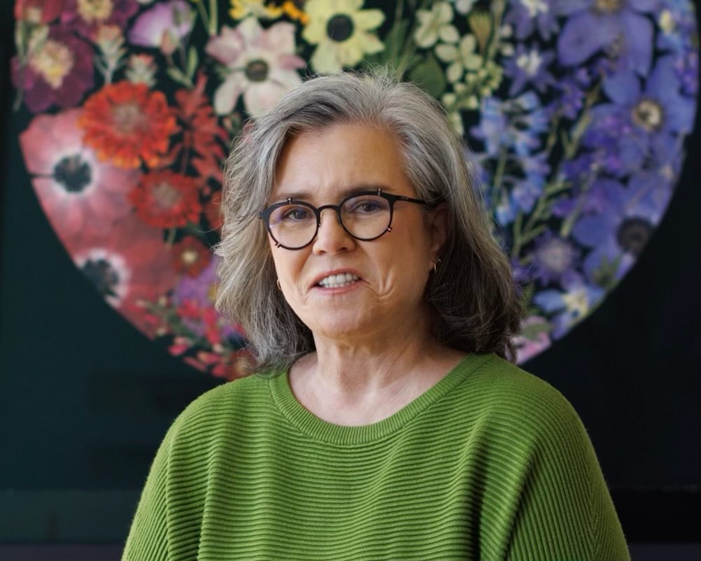 Rosie O'Donnell poses in front of a colorful floral artwork while wearing a green sweater and round glasses, looking calmly toward the camera in a softly lit setting. | Source: Instagram/rosie