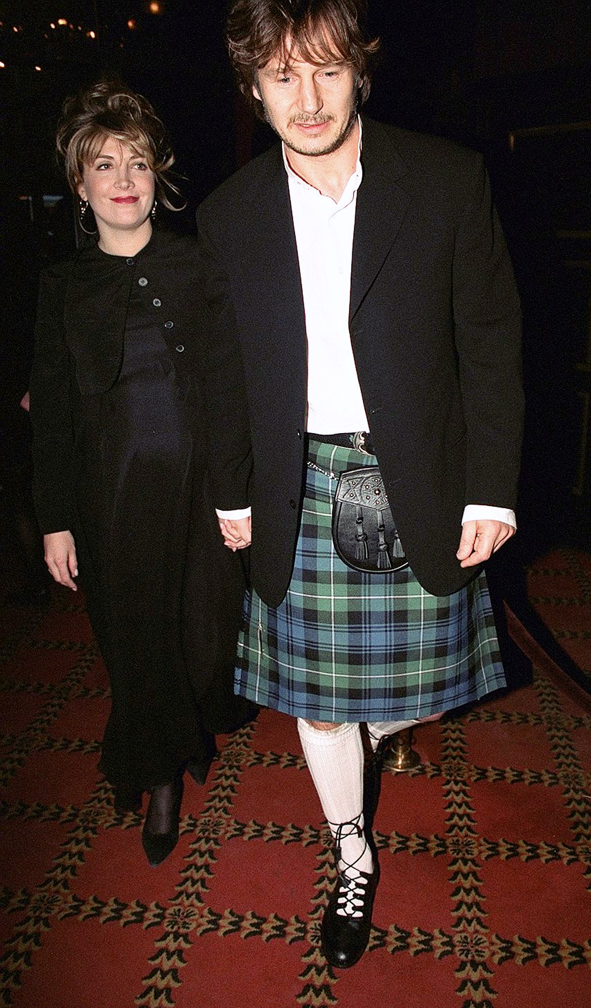 Liam Neeson, wearing a traditional kilt, holds hands with Natasha Richardson as they arrive at the premiere of “Rob Roy” at the Ziegfeld Theatre in April 1995. The couple walks together on the red carpet, marking the release of Neeson’s film in which he played the title role.