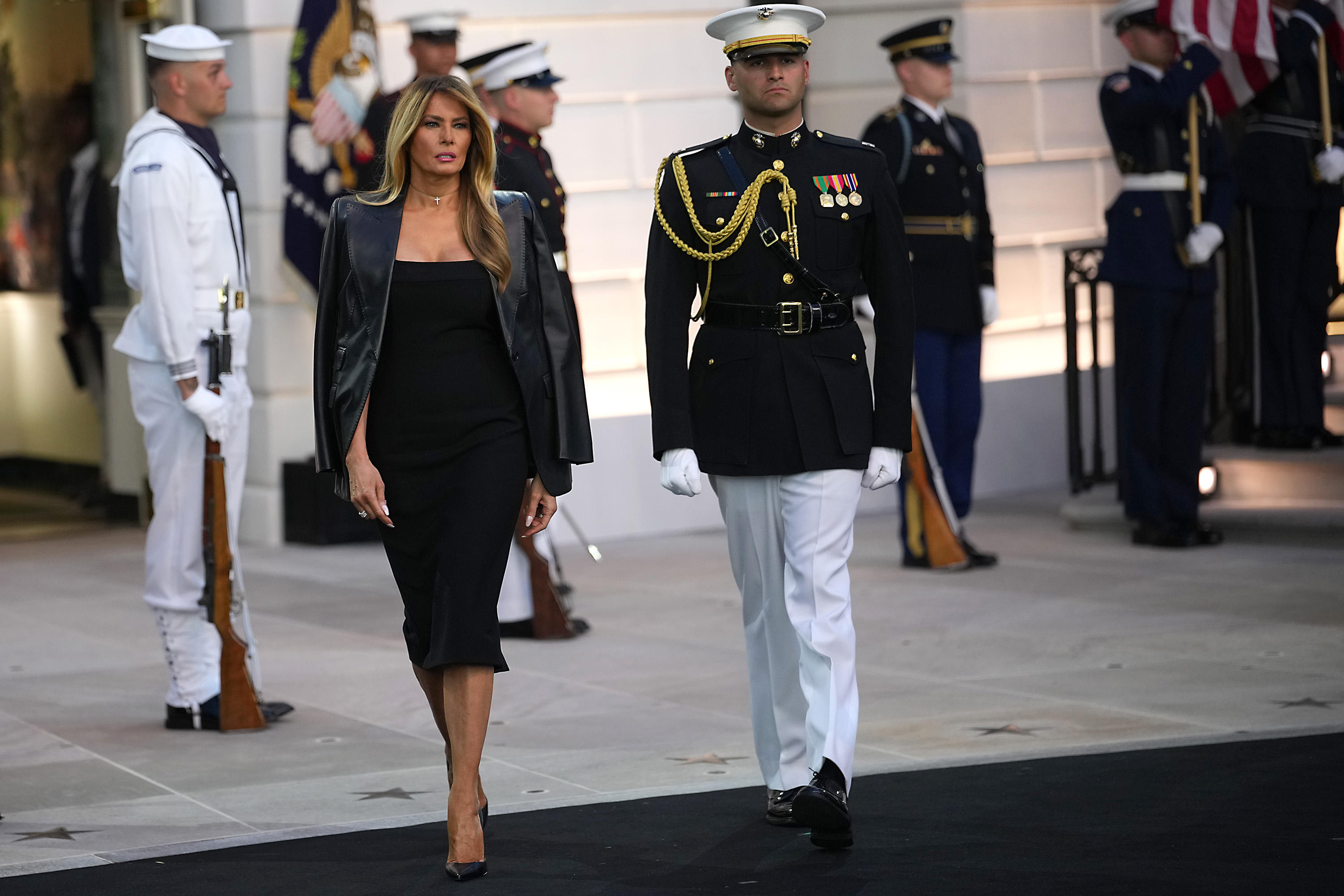 Melania Trump steps across the South Lawn in a fitted black dress and draped leather jacket. Her long highlighted hair, defined cheekbones, and sculpted facial features draw attention.