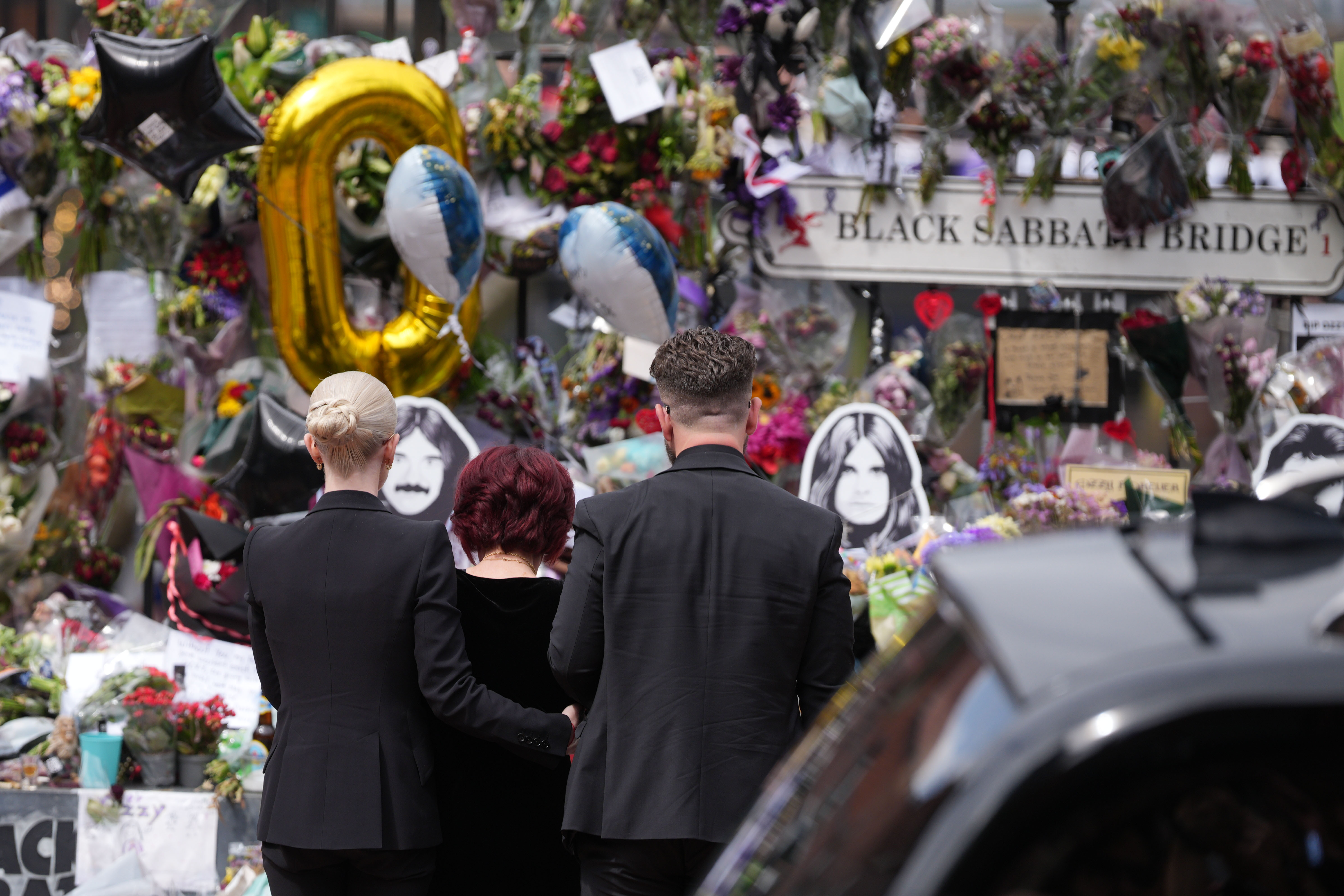 Kelly Osbourne, Sharon Osbourne and Jack Osbourne stop to view tributes to the late Ozzy Osbourne from fans at Black Sabbath Bench and Bridge as his funeral cortege travels through his home city of Birmingham on July 30, 2025 in Birmingham, England. | Source: Getty Images