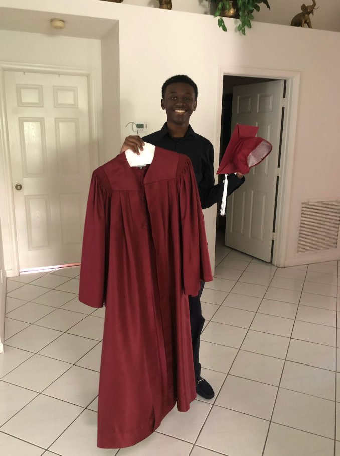 Smiling proudly indoors, Donovan Metayer, holds up his maroon graduation gown alongside his matching cap, marking a milestone moment filled with anticipation and achievement. | Source: Gofundme